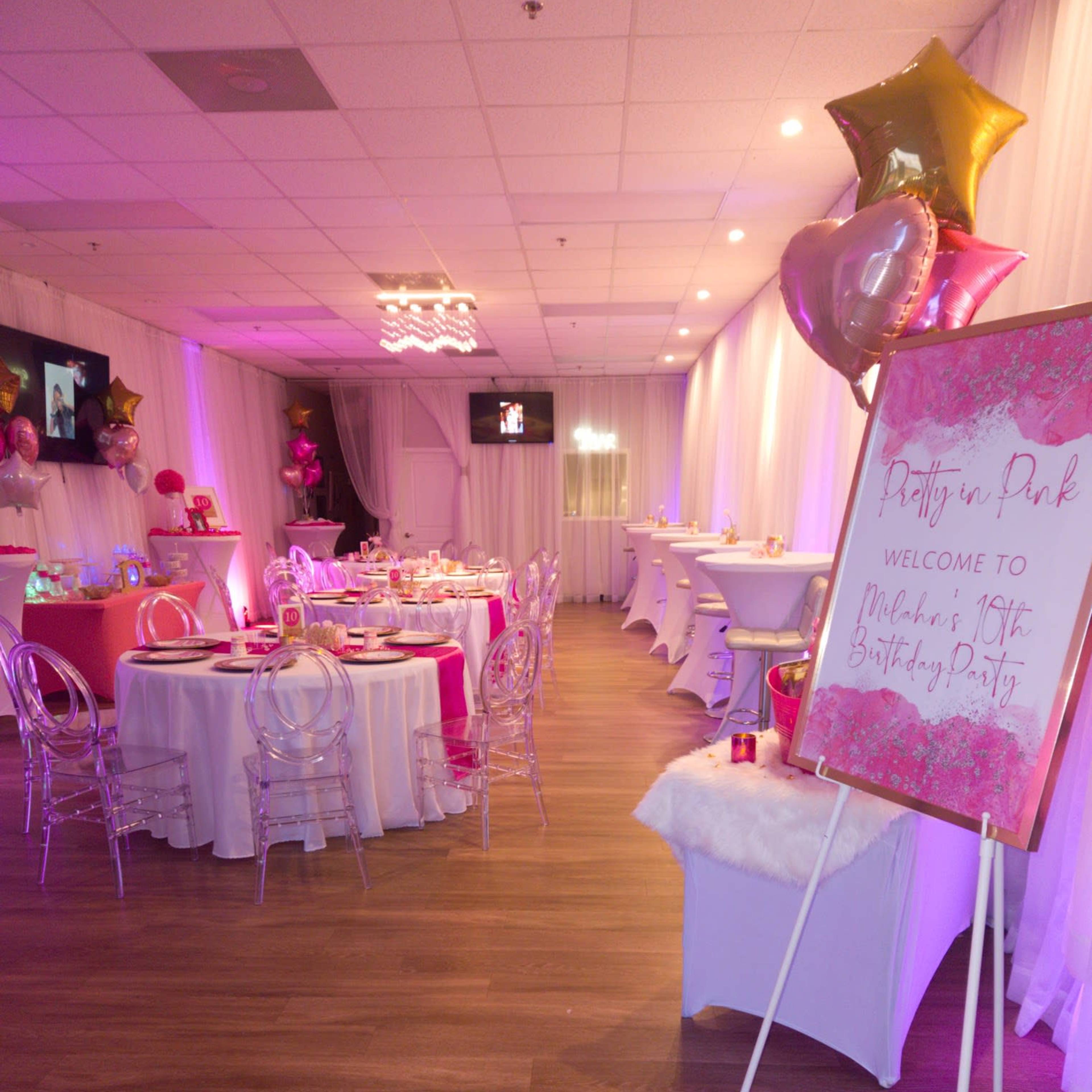 A brightly decorated indoor party space with tables set for a birthday celebration, featuring pink accents, balloons, and a welcoming sign.