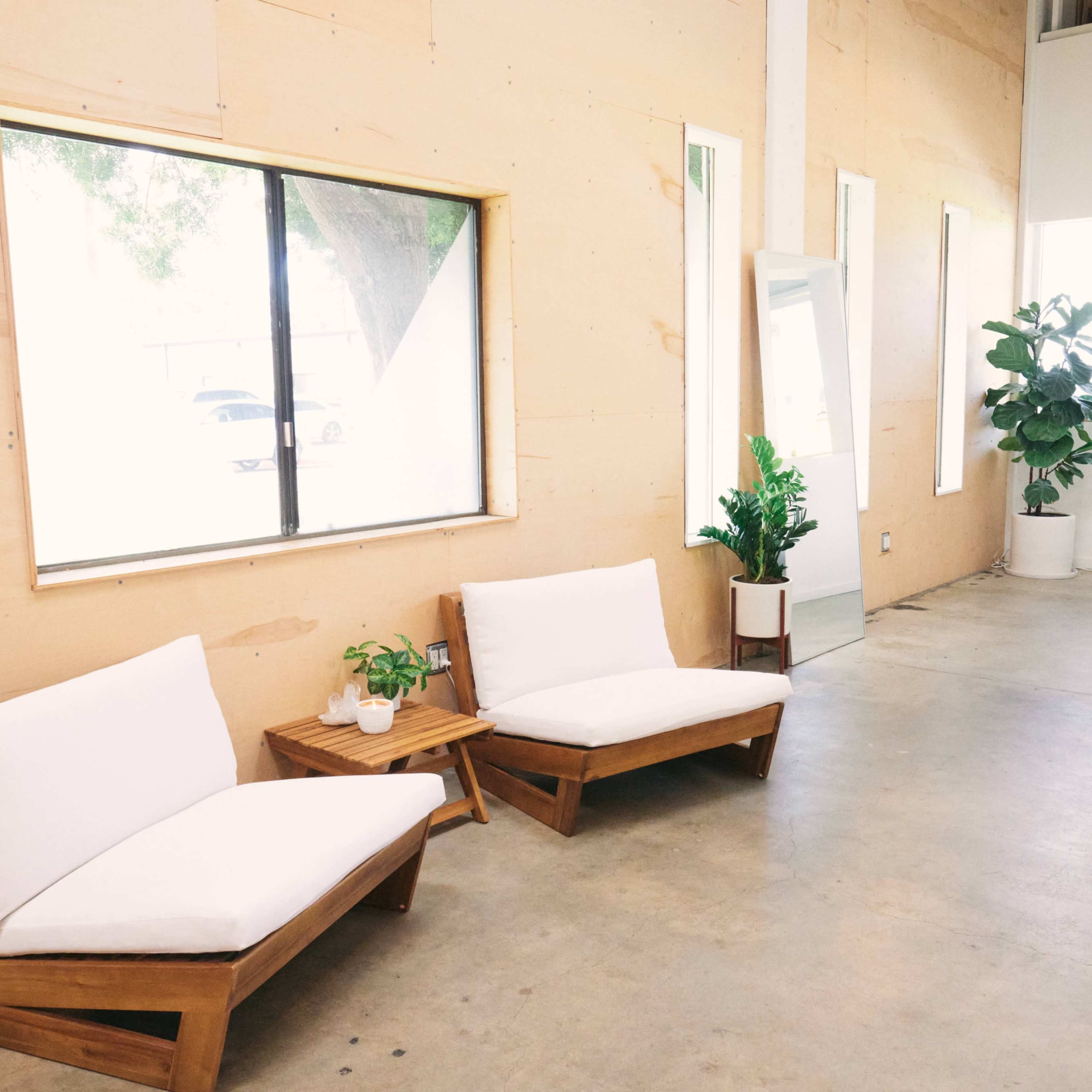 A bright, minimalist interior featuring two wooden chairs with white cushions, a plant in a pot, and large windows providing natural light.