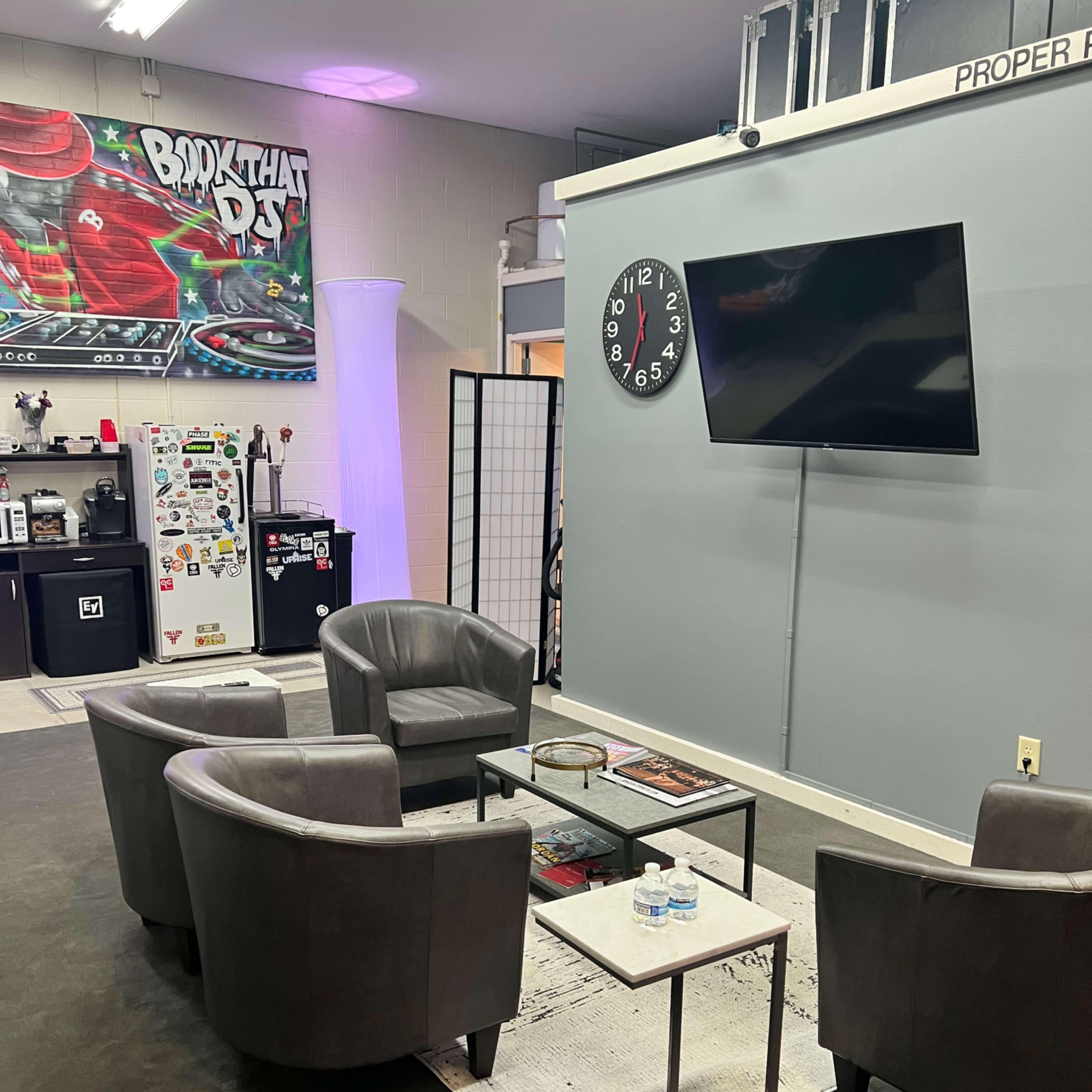 A seating area with four gray chairs and a small table is set against a wall featuring a large clock and a TV in a mixed-use space decorated with a colorful mural.