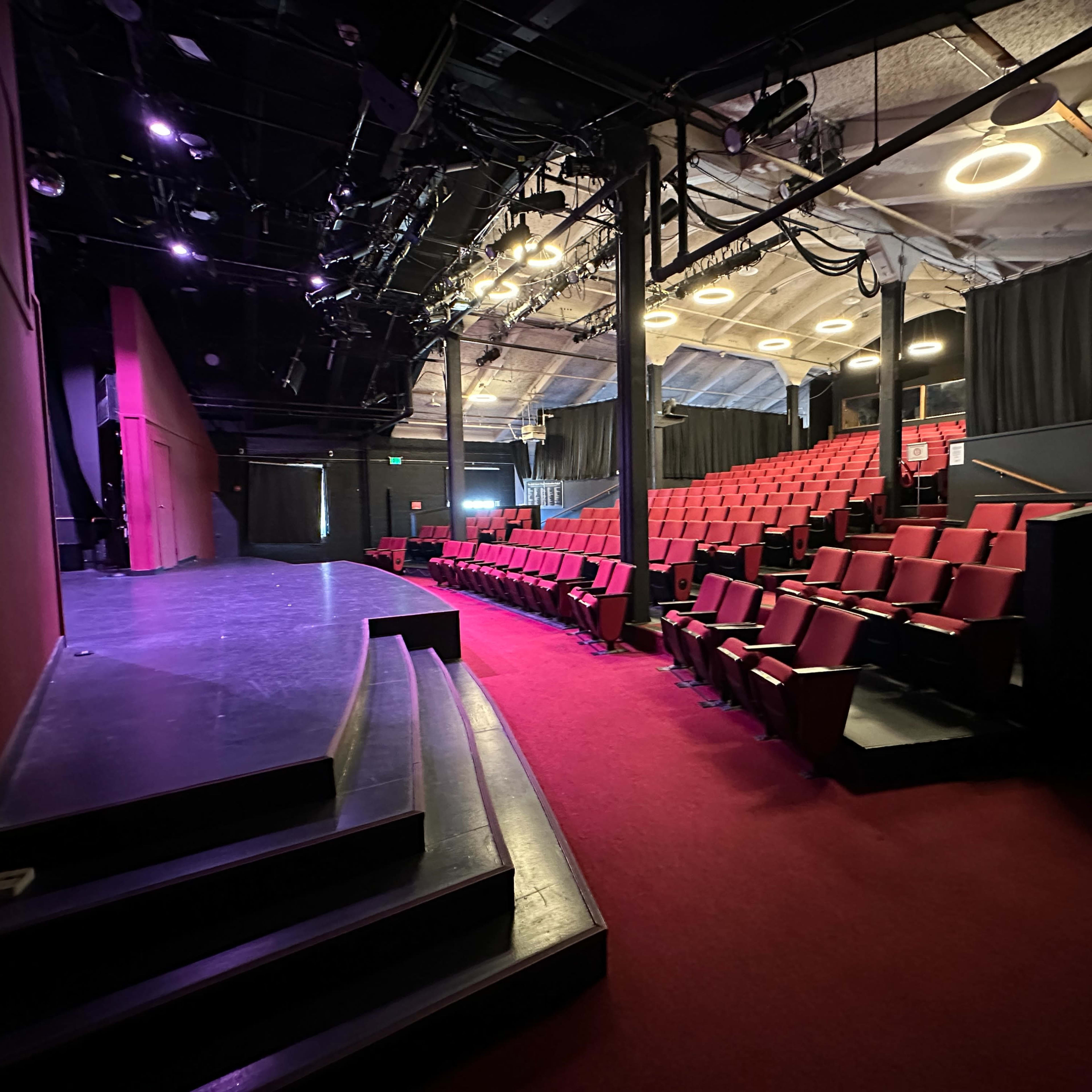 The interior of a theater with a stage, red seating, and dimly lit surroundings.