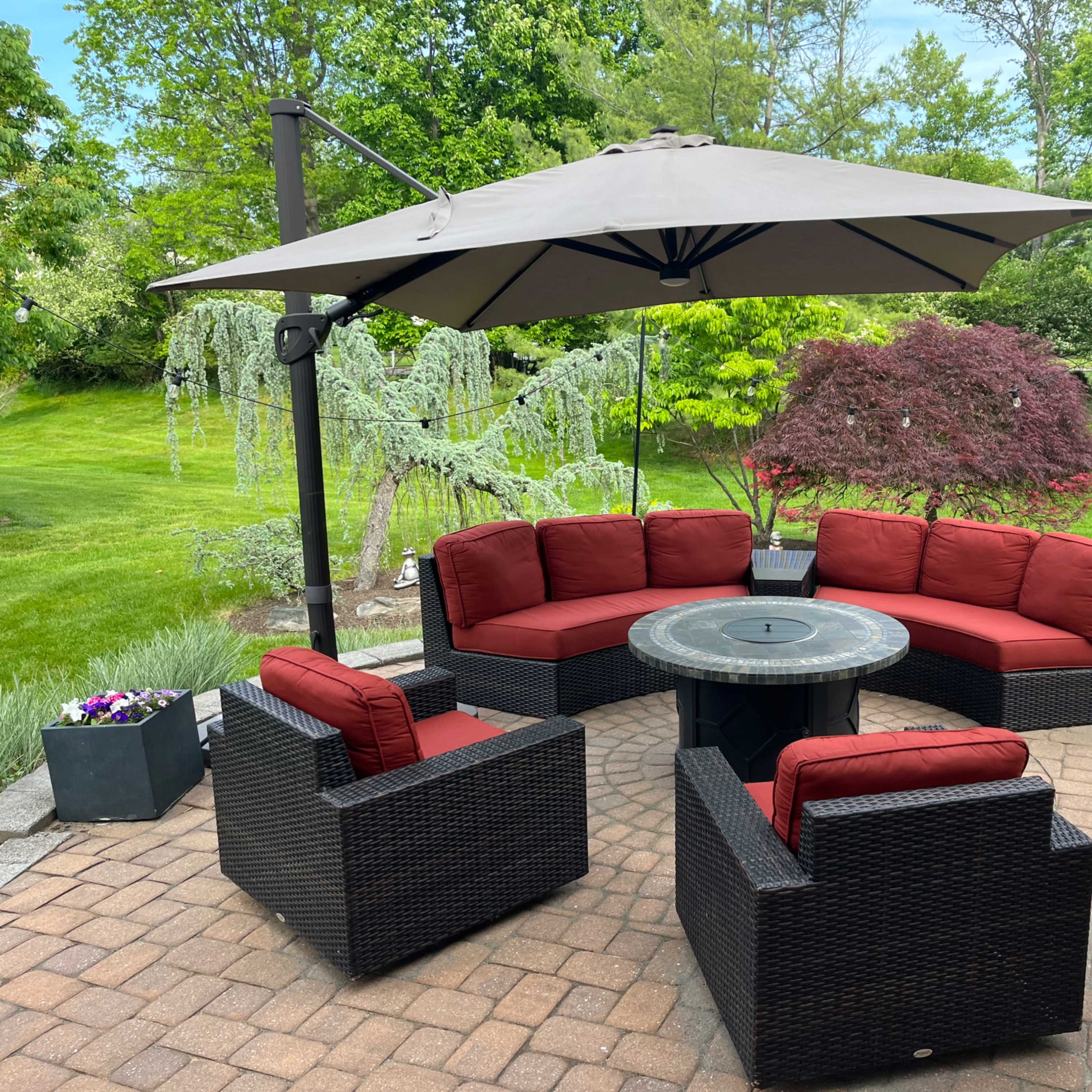 A circular outdoor seating arrangement with red cushions is placed under a large umbrella on a stone patio surrounded by green grass and trees.