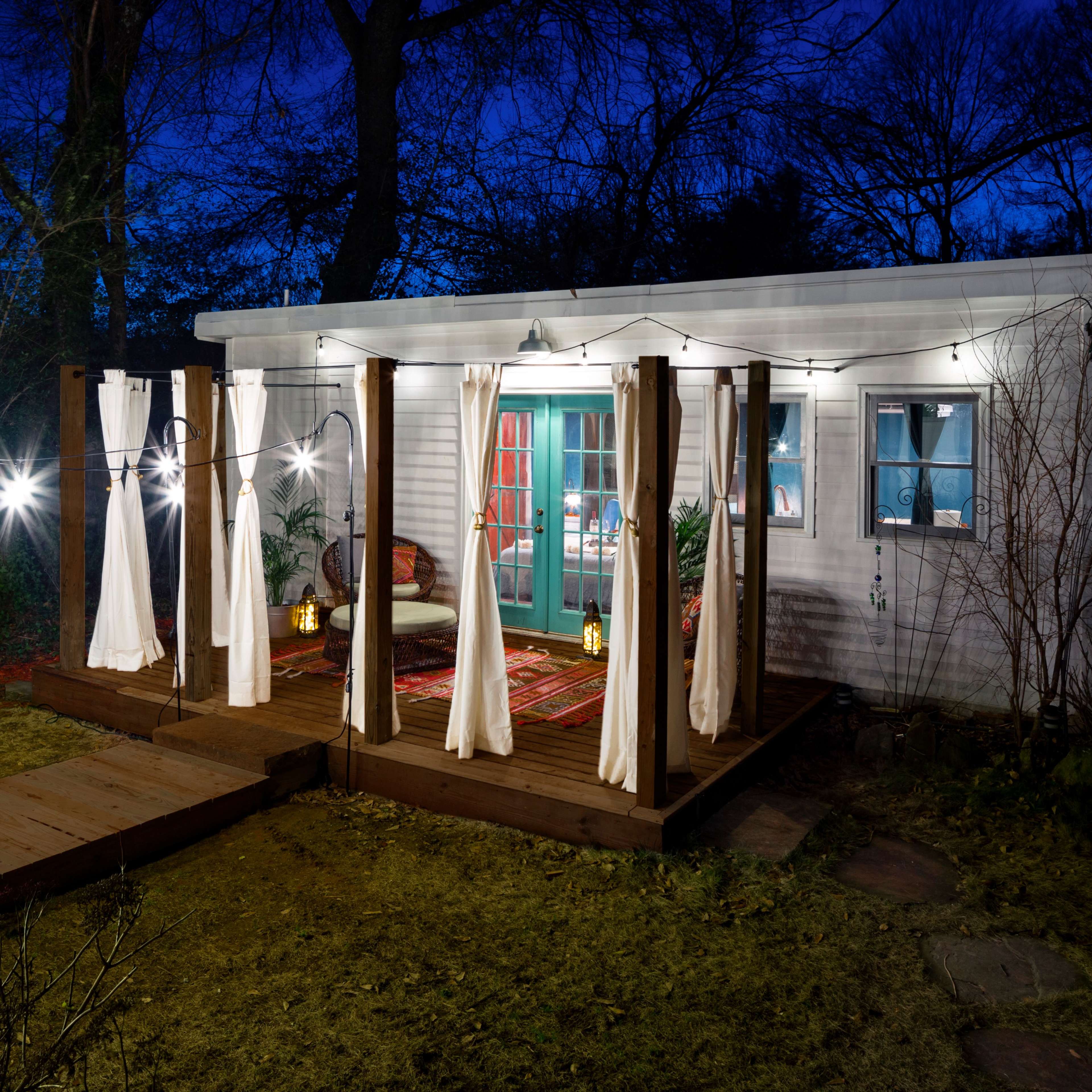A cozy outdoor patio features a small wooden deck with curtains, lanterns, and string lights, adjacent to a well-lit cabin at dusk.