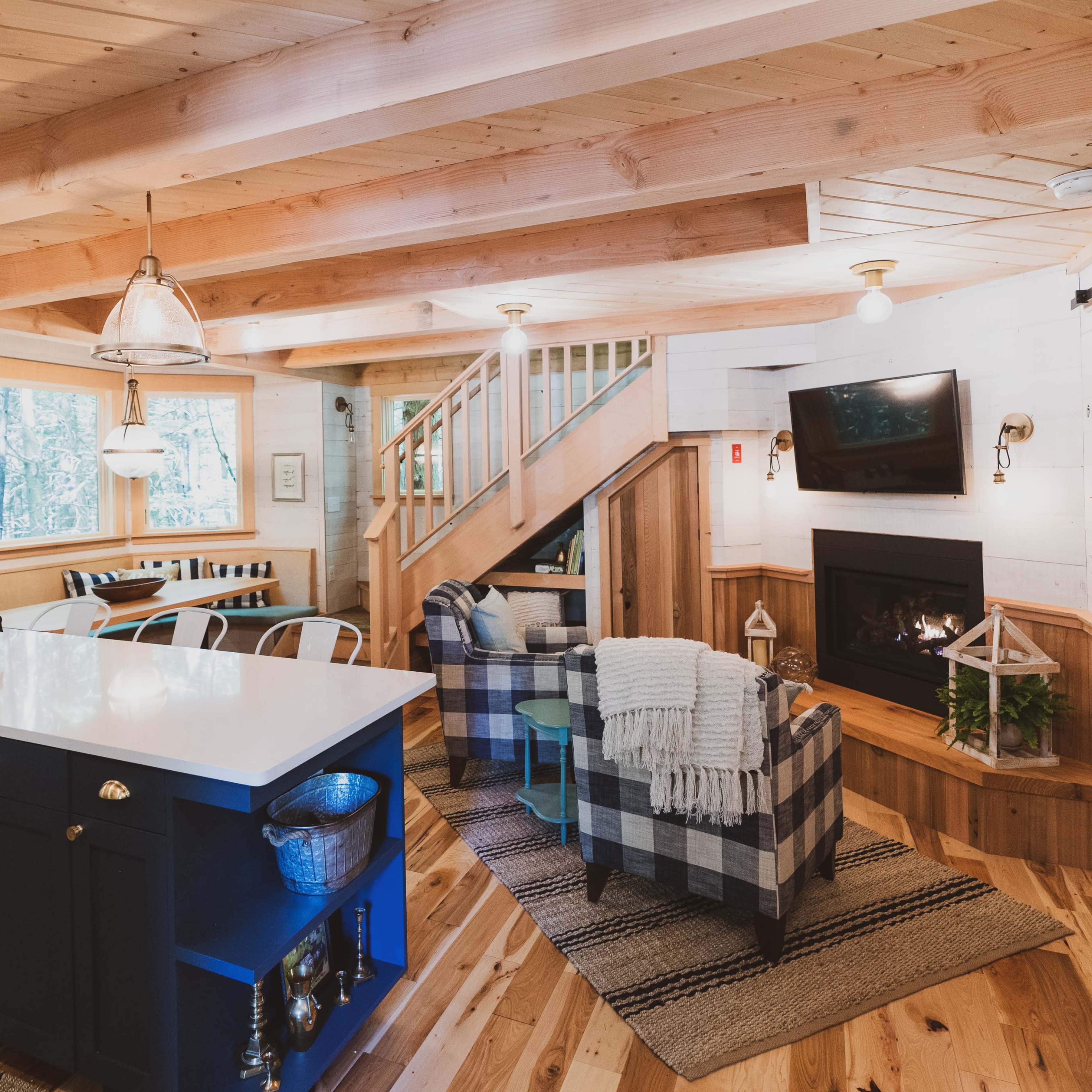 A cozy, modern cabin interior featuring a living area with wooden beams, a fireplace, a kitchen island with a sink, and stylish seating arrangements.
