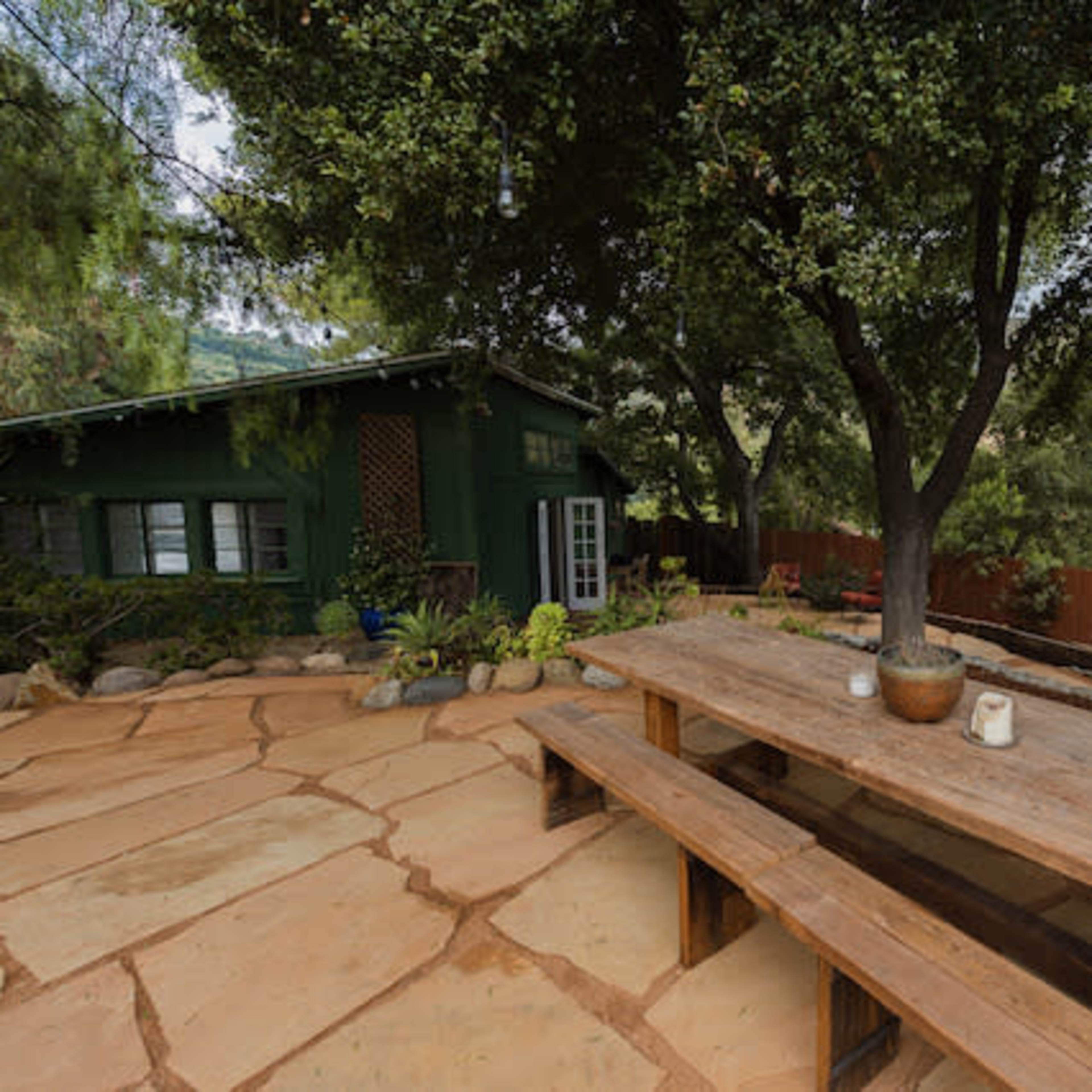 A large wooden table sits on a stone patio surrounded by trees, with a green building in the background.