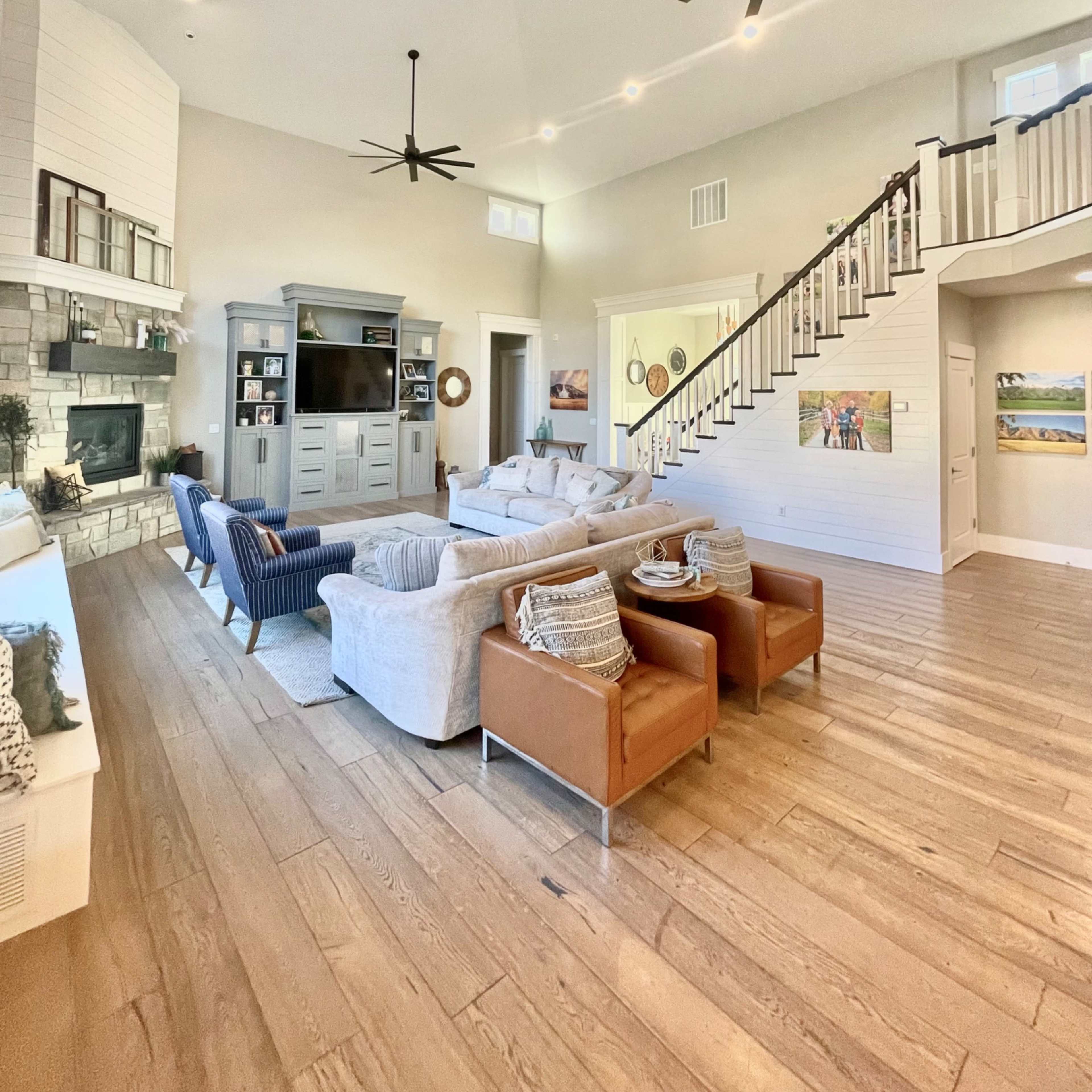 A spacious living room with a large sectional sofa, two armchairs, and a television set against a light-colored wall, illuminated by several ceiling fans and recessed lighting.