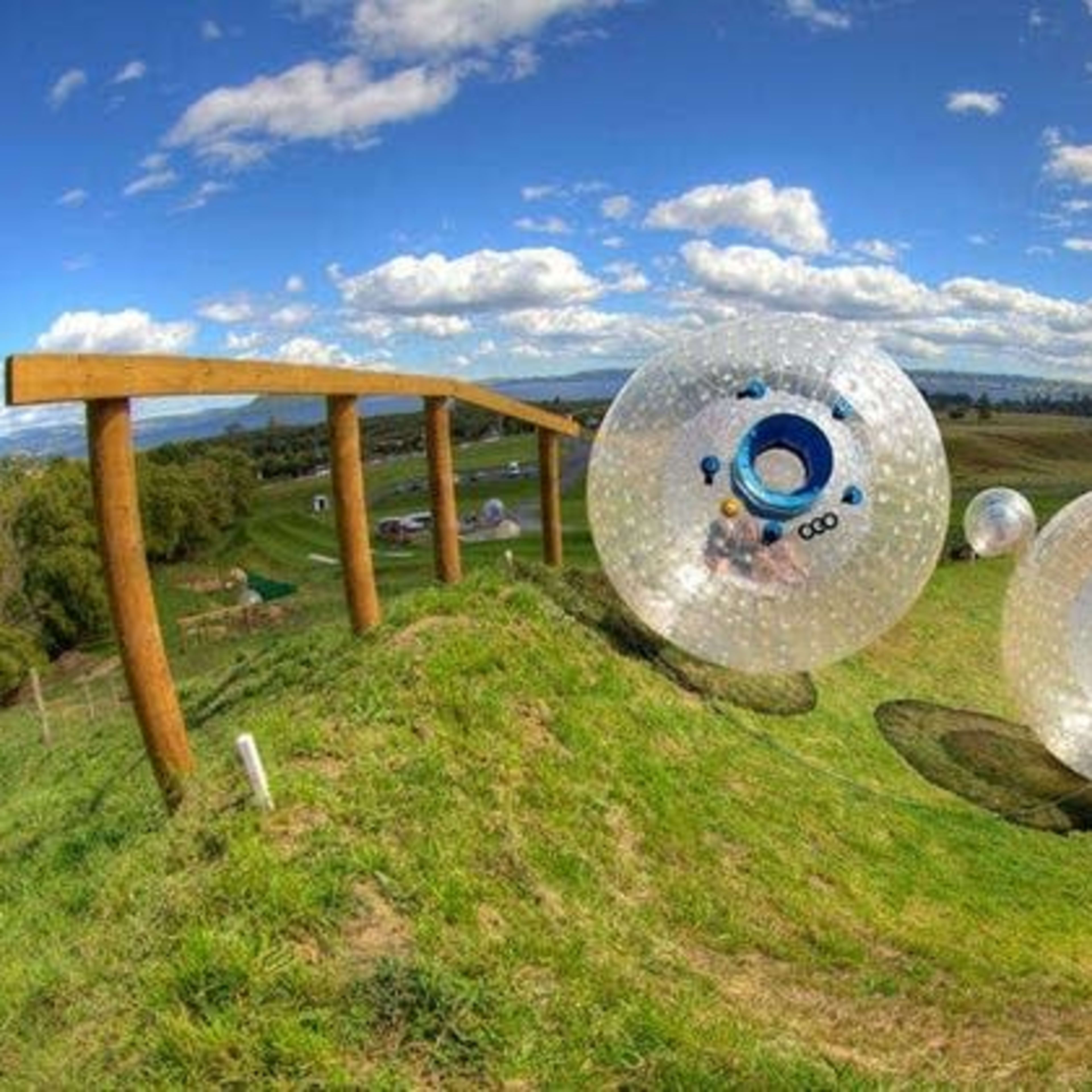 Transparent spheres roll down a grassy hill along a wooden track under a blue sky with clouds.