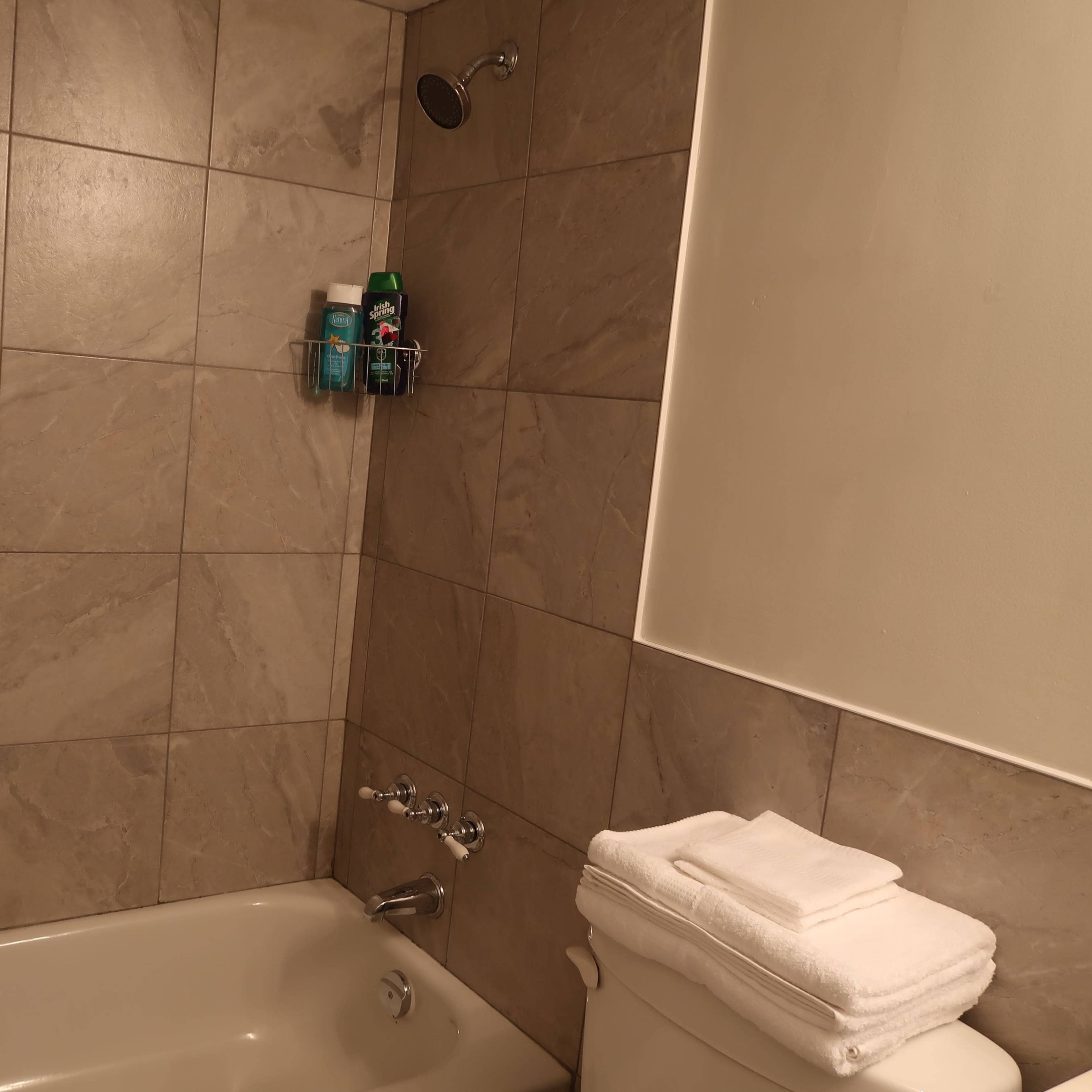A bathroom with a bathtub, a showerhead, a wall-mounted shelf with toiletries, and neatly placed towels on the toilet tank.