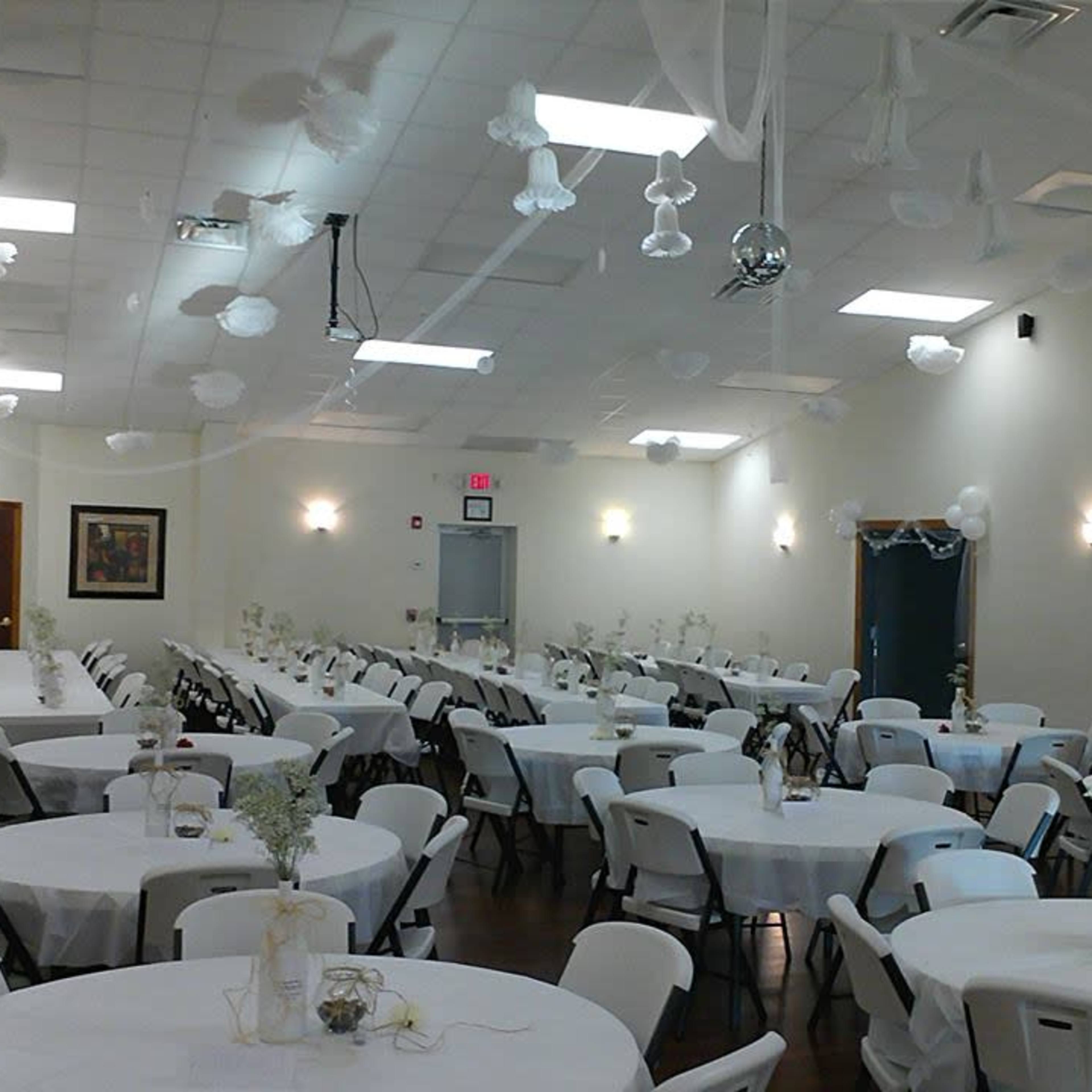A spacious banquet hall featuring round tables covered with white tablecloths and decorated with floral arrangements, with a disco ball and white decorations hanging from the ceiling.