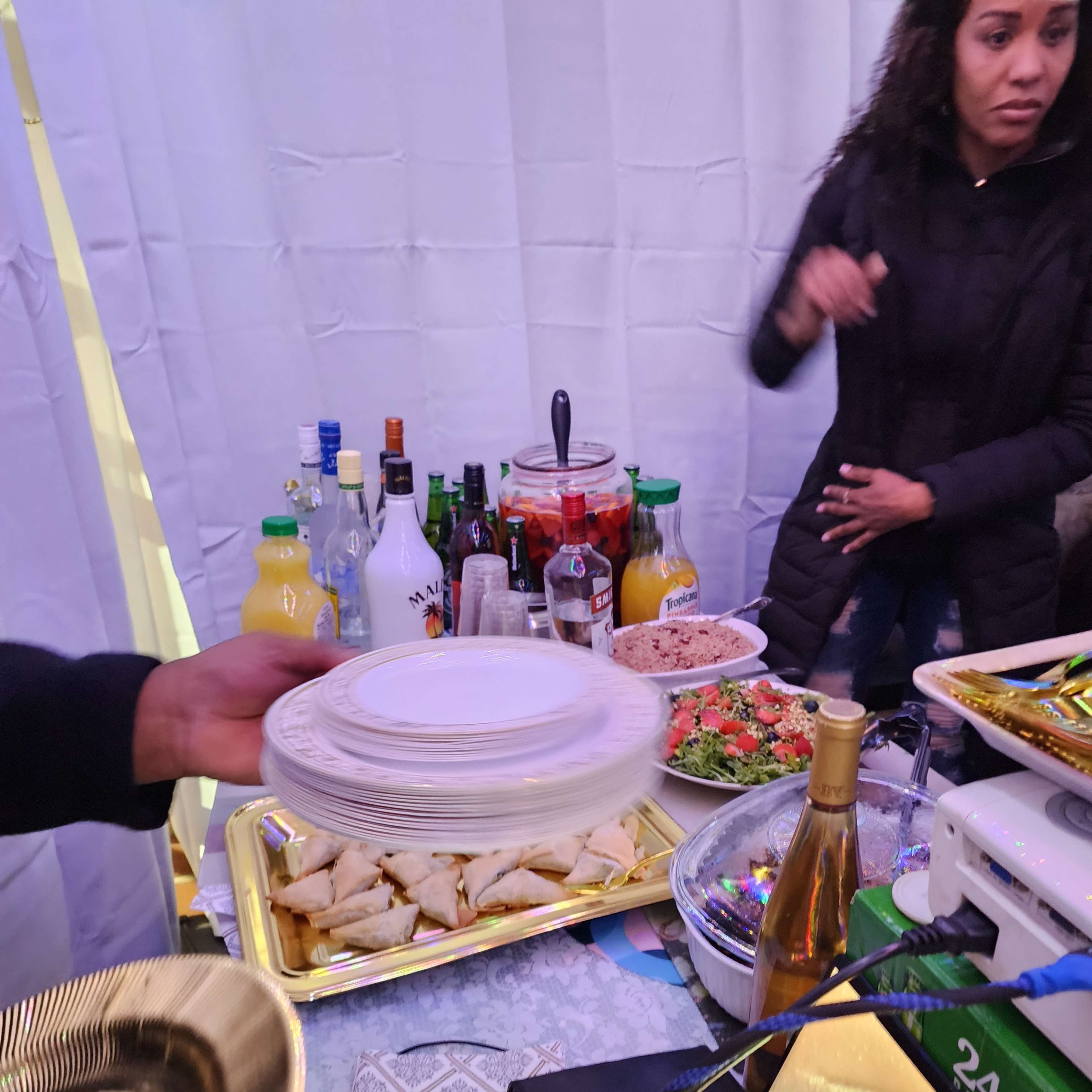 A table is set with various bottles, dishes of food, and a person holding a stack of plates in a gathering space.