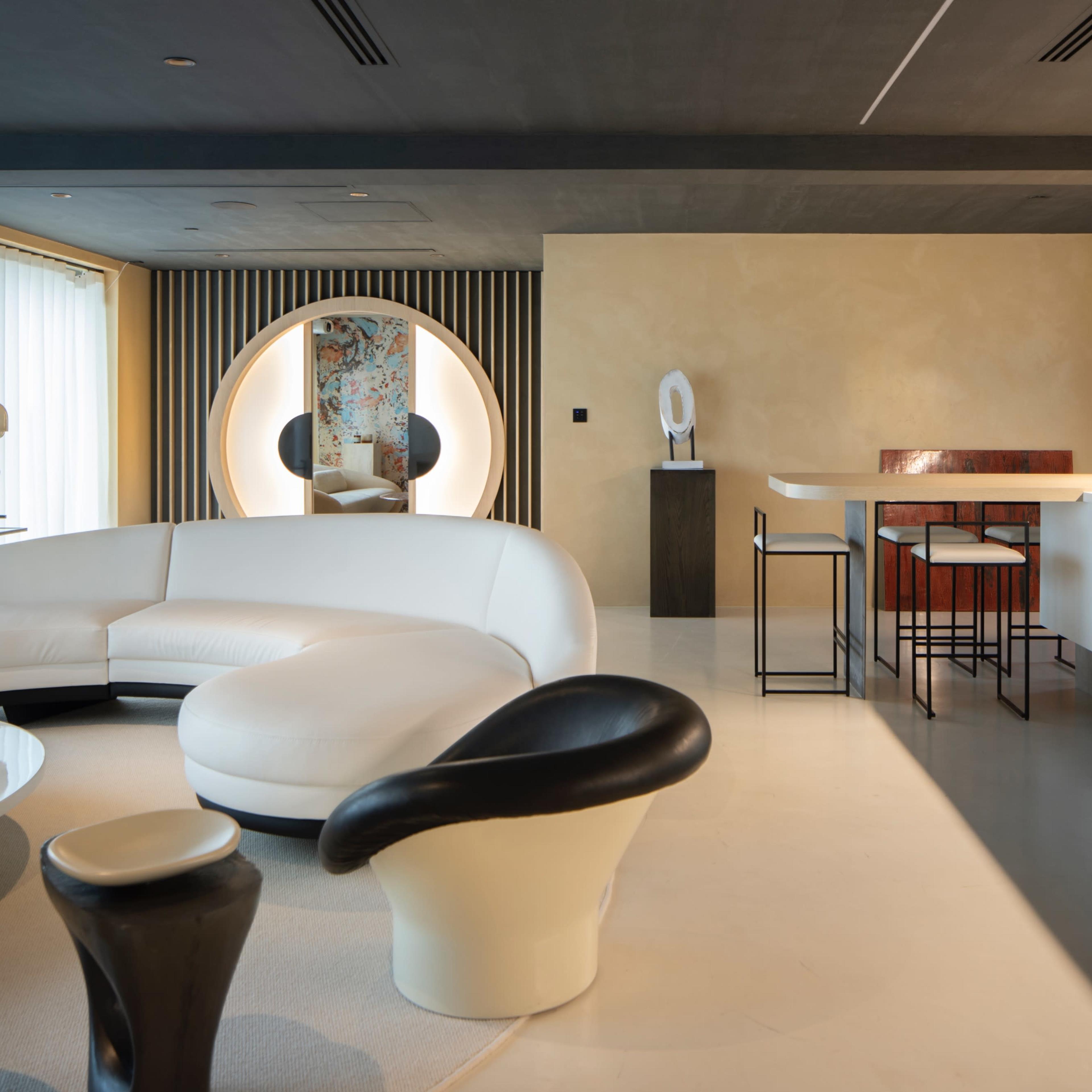 A modern interior space featuring a white curved sofa, a round mirror, a minimalist bar area with high stools, and a light-colored floor.