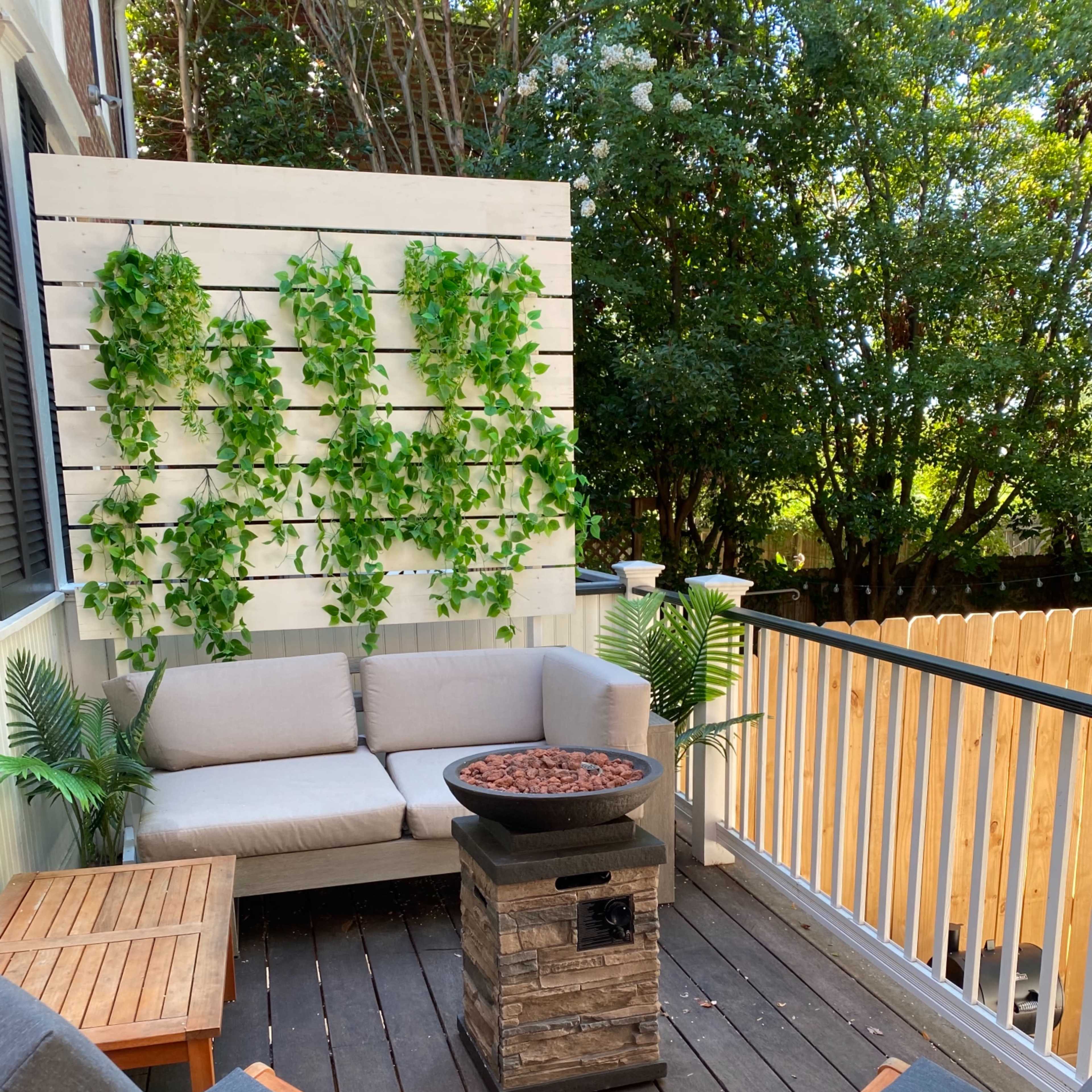 A cozy outdoor seating area with a gray couch and two wooden chairs arranged around a stone fire pit, surrounded by decorative greenery and wooden fencing.