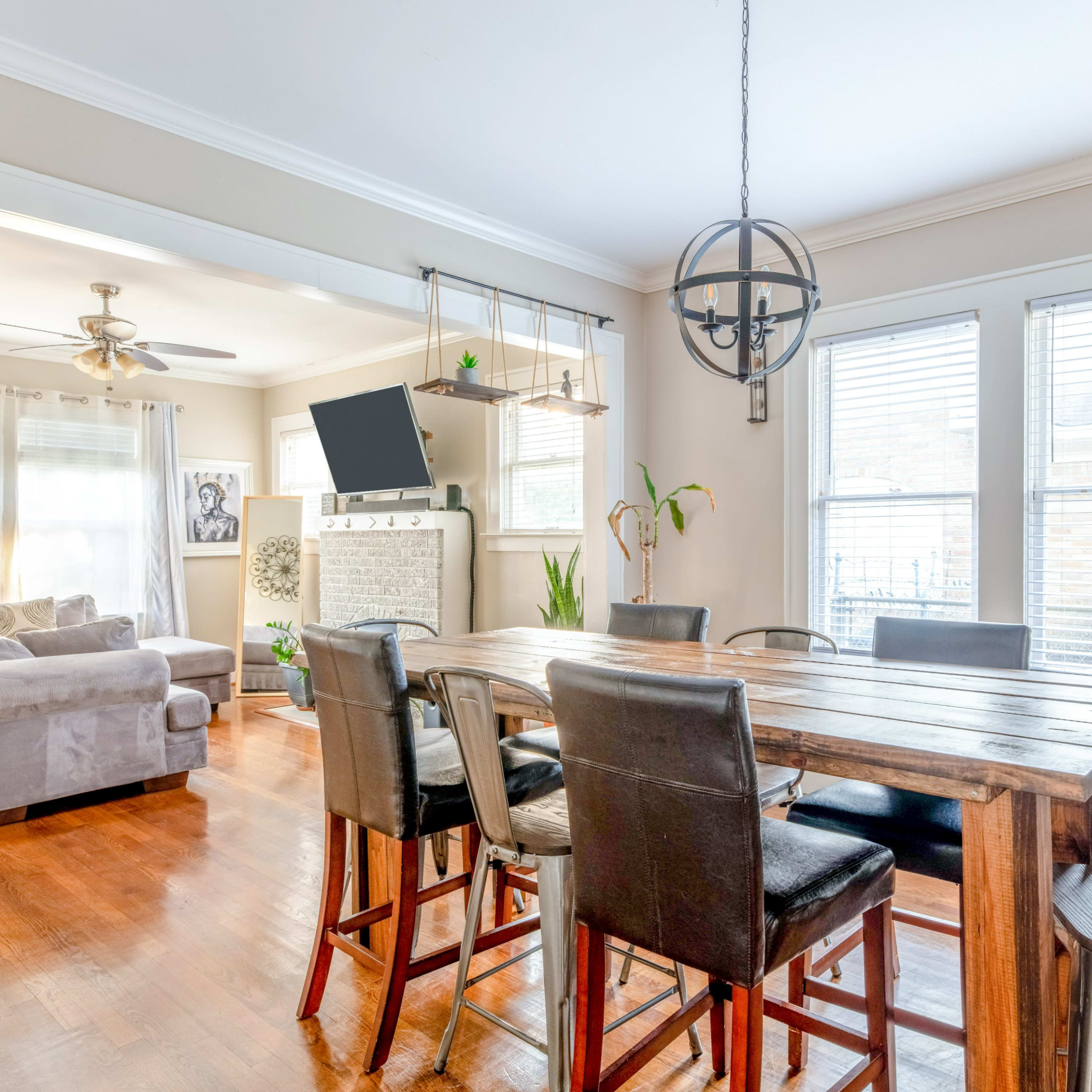 A wooden dining table with black chairs is set in a bright room featuring large windows, a comfortable couch, and a wall-mounted TV.