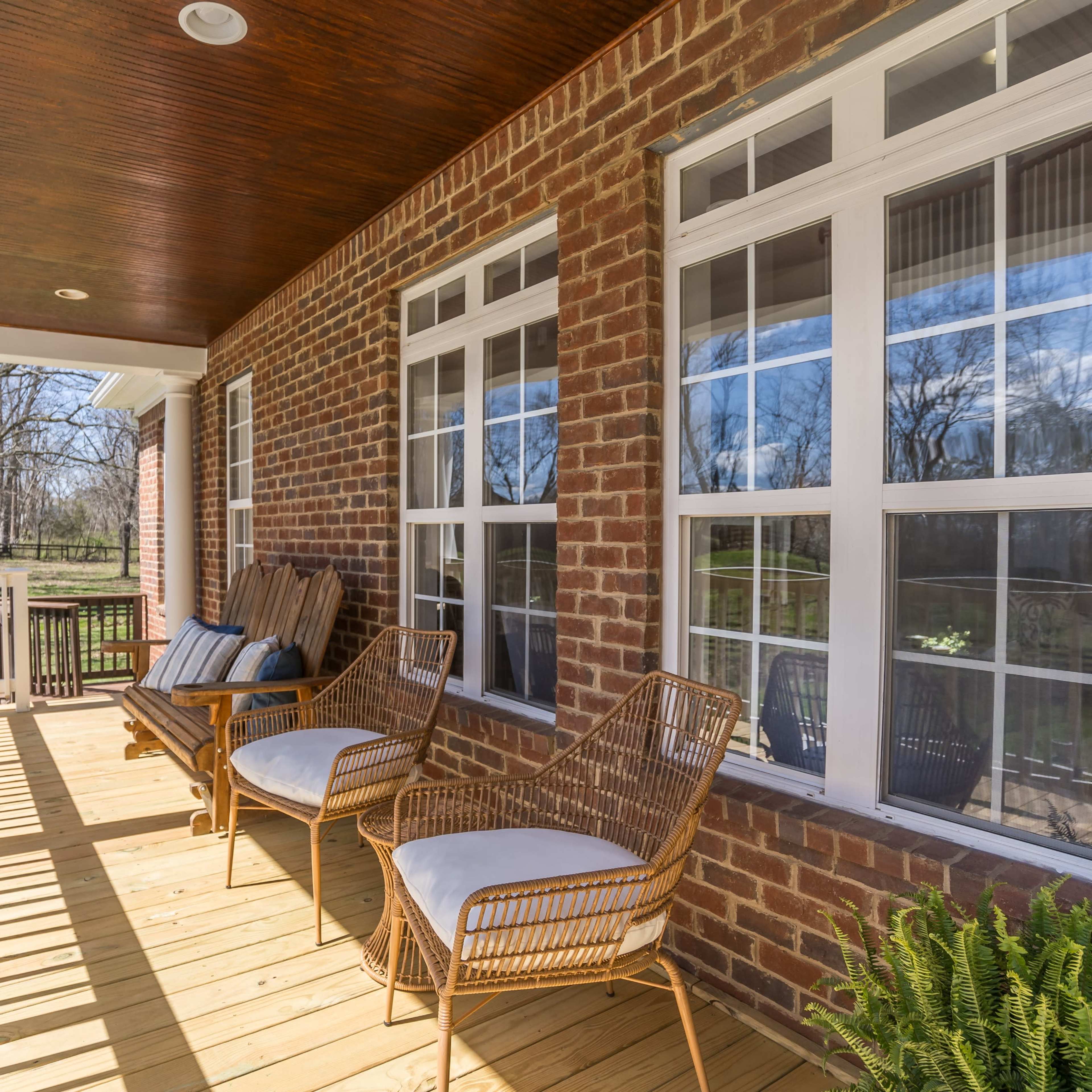 A porch with wooden flooring, two wicker chairs, and large windows set in a brick wall, overlooking a wooded area.