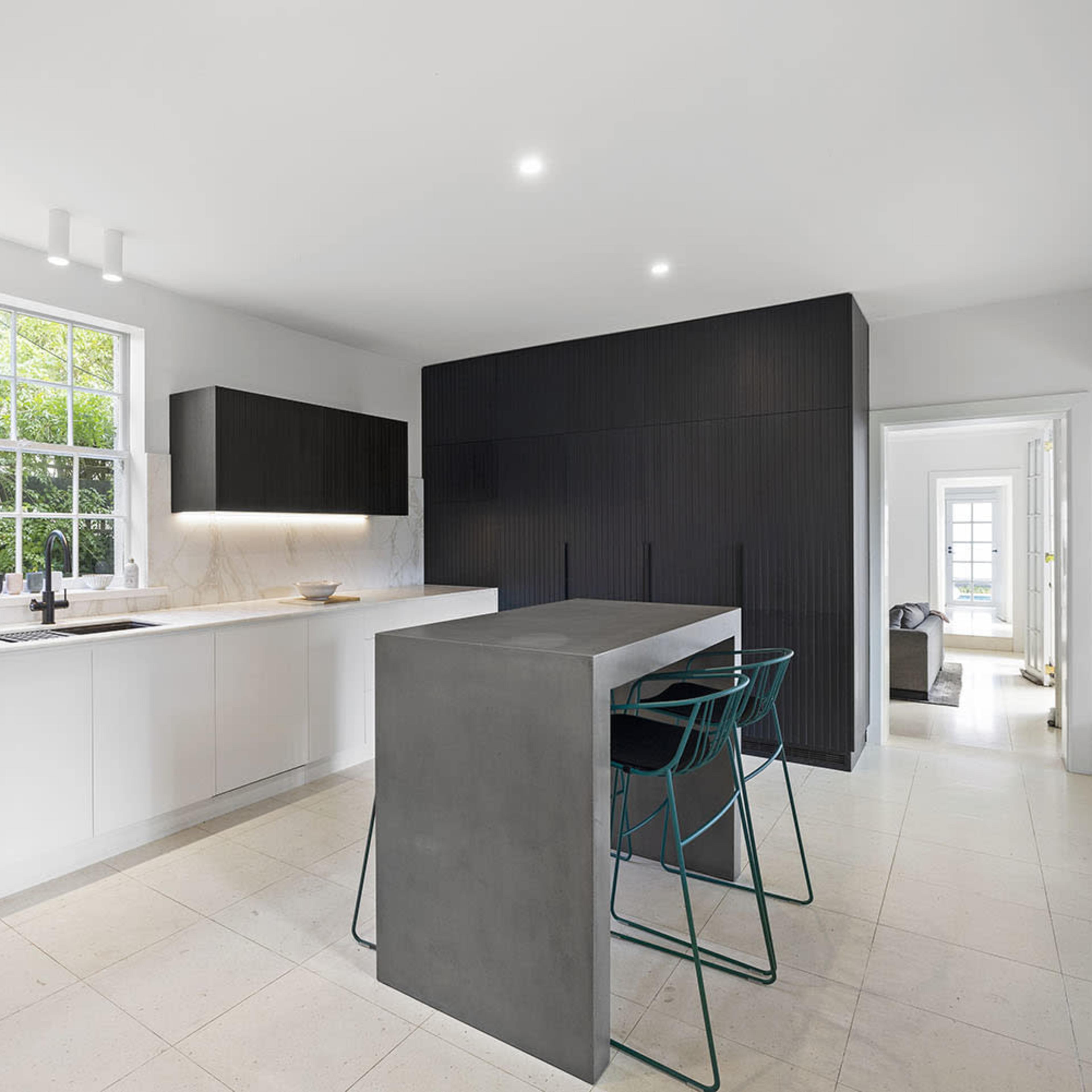 A modern kitchen featuring a minimalist design with white cabinetry, a dark wood island, and large windows bringing in natural light.