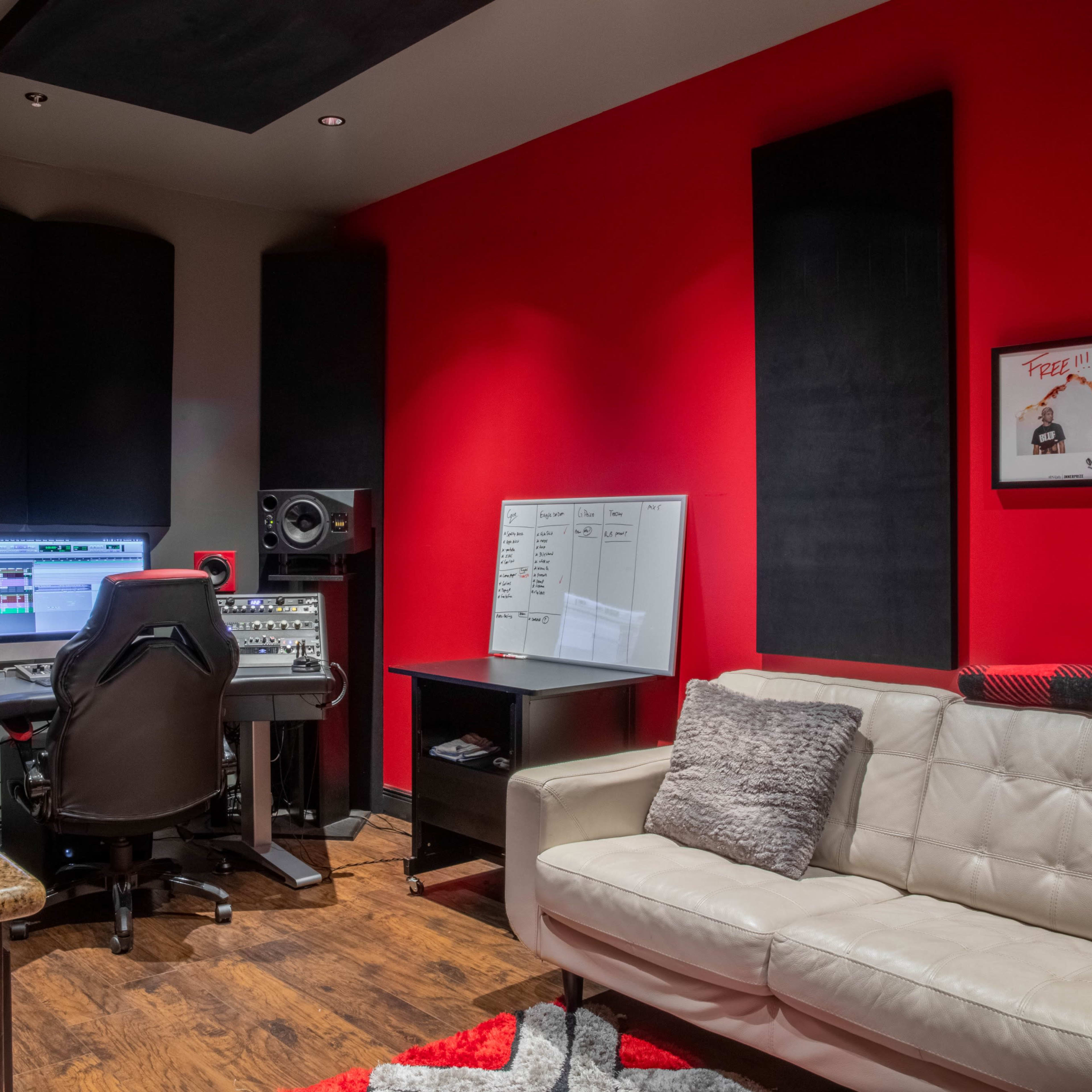 A modern recording studio featuring a central workstation with speakers, a computer monitor, and a comfortable white couch against a backdrop of red and black walls.