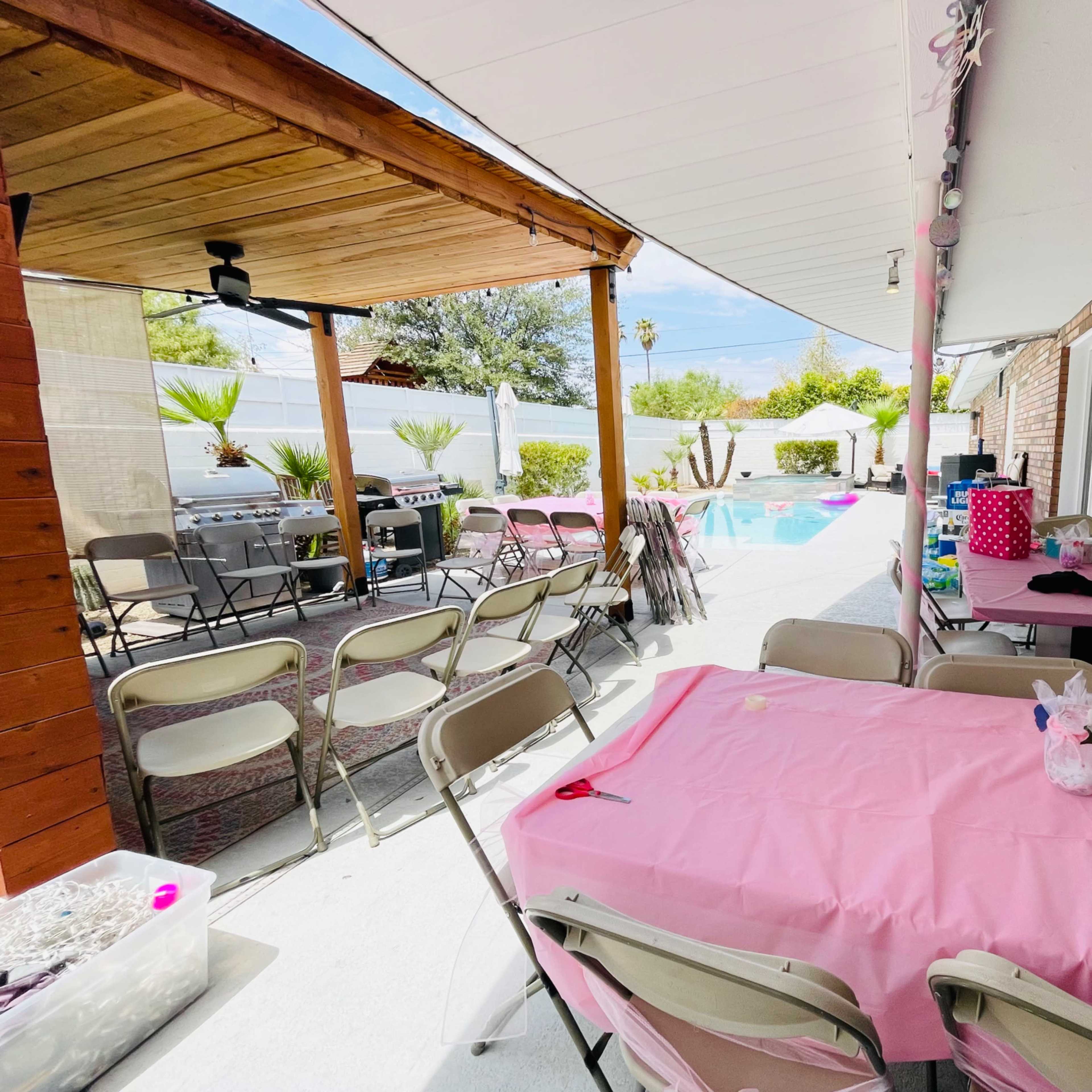 A backyard setting with a covered patio area, tables covered in pink cloth, and chairs arranged around a swimming pool.
