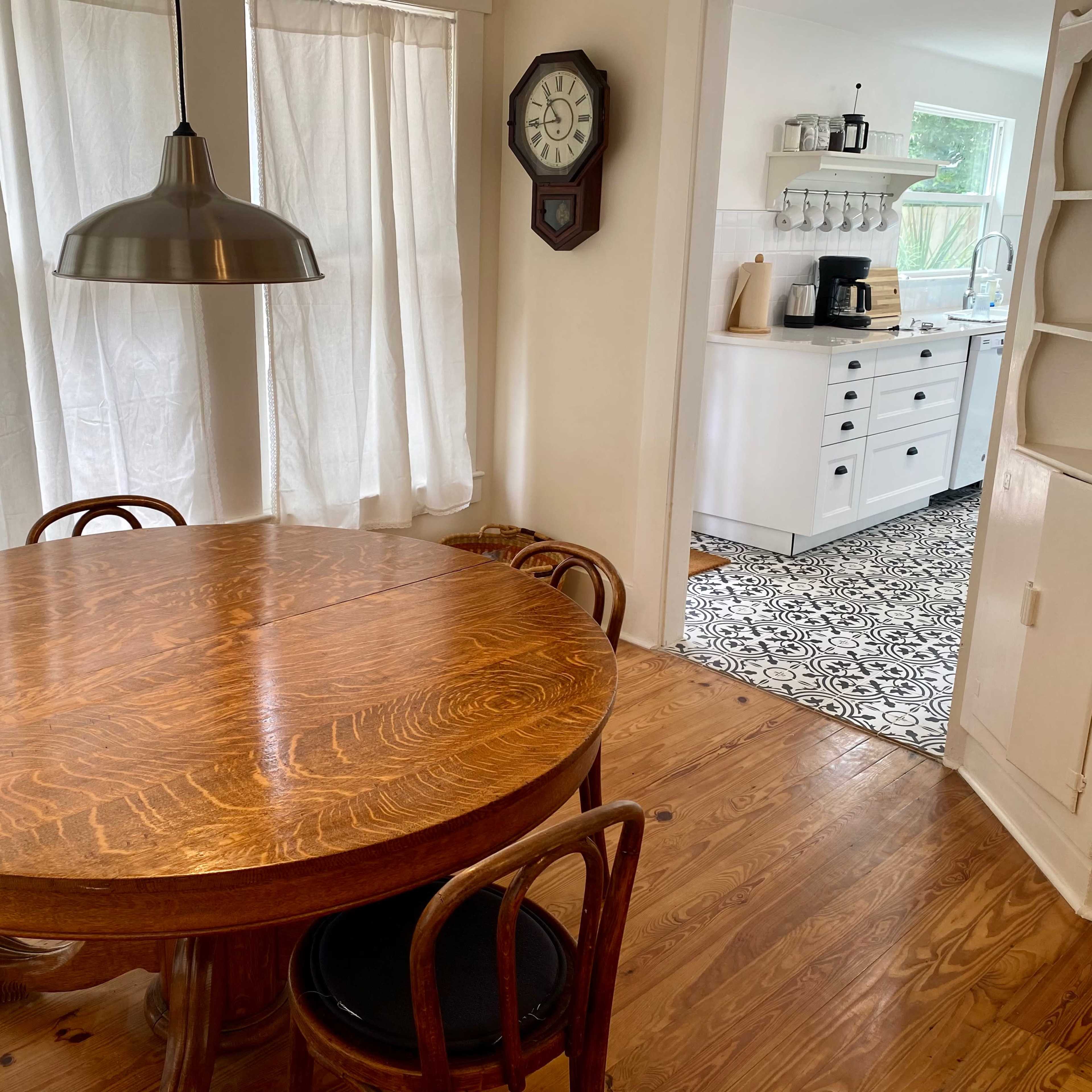 A wooden dining table with four chairs is positioned near a window, leading to a kitchen visible in the background.