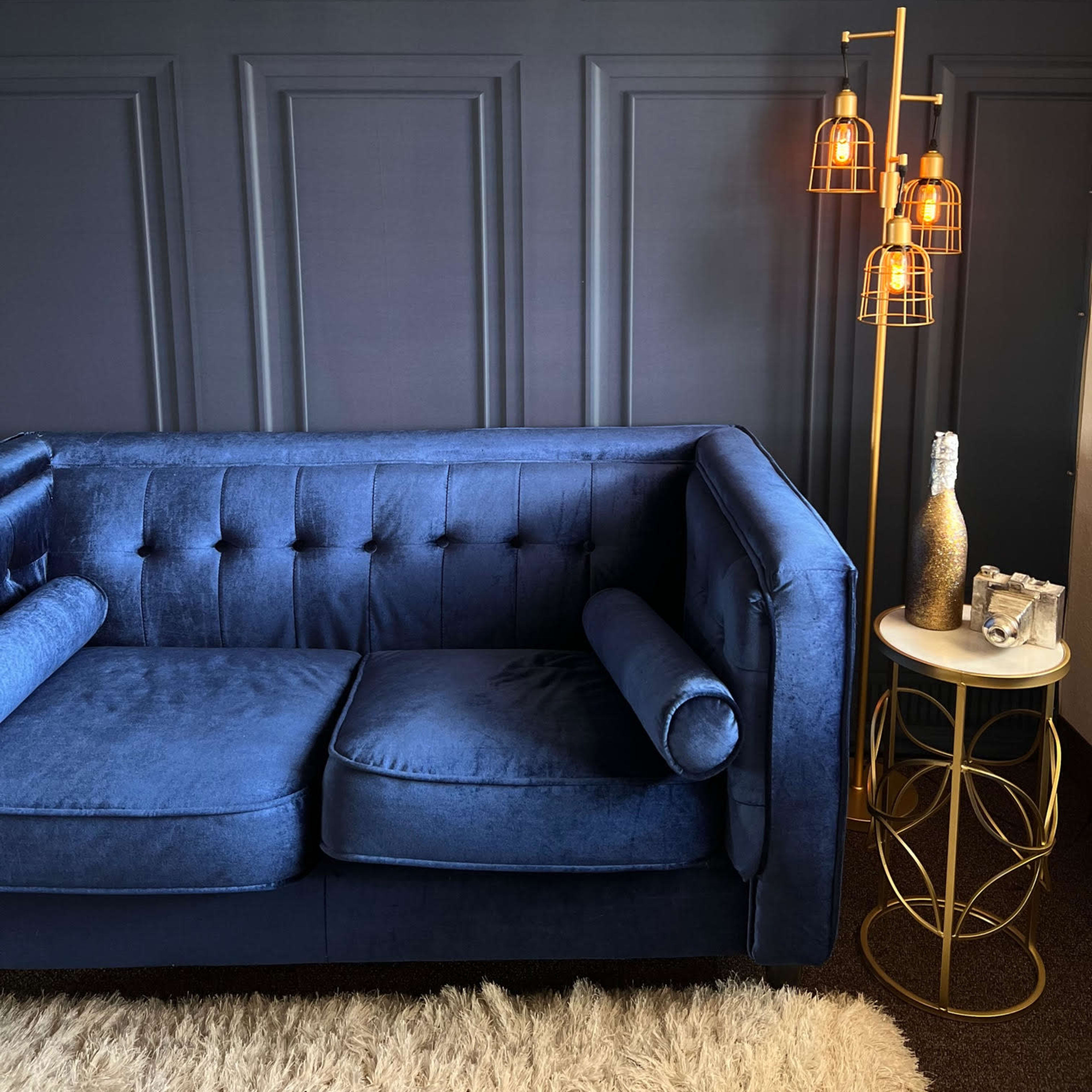 A blue velvet couch against a dark paneled wall, accompanied by a gold lamp and a circular side table.