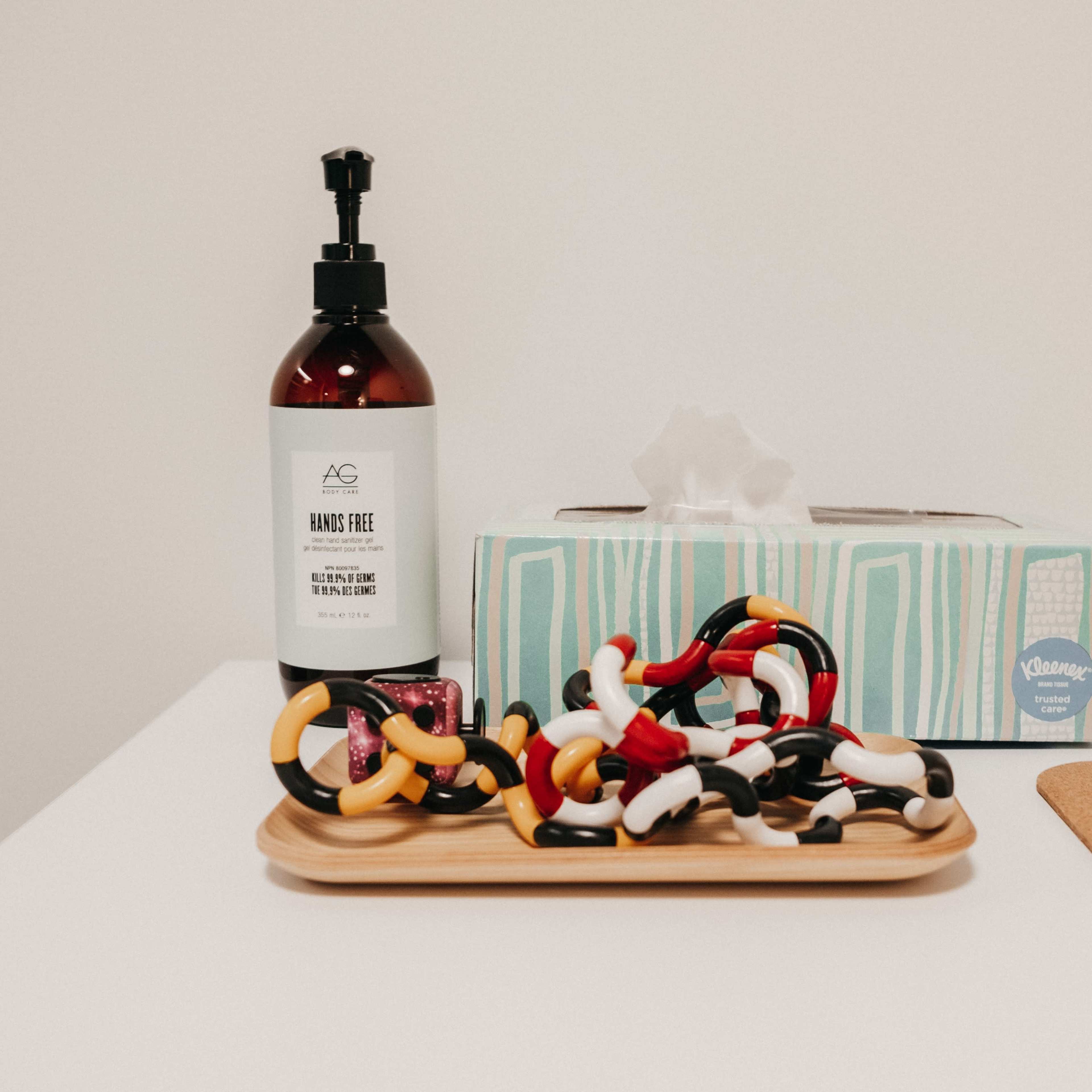 A bottle of hand sanitizer, a tissue box, and a tray of colorful hair ties are arranged on a white table.