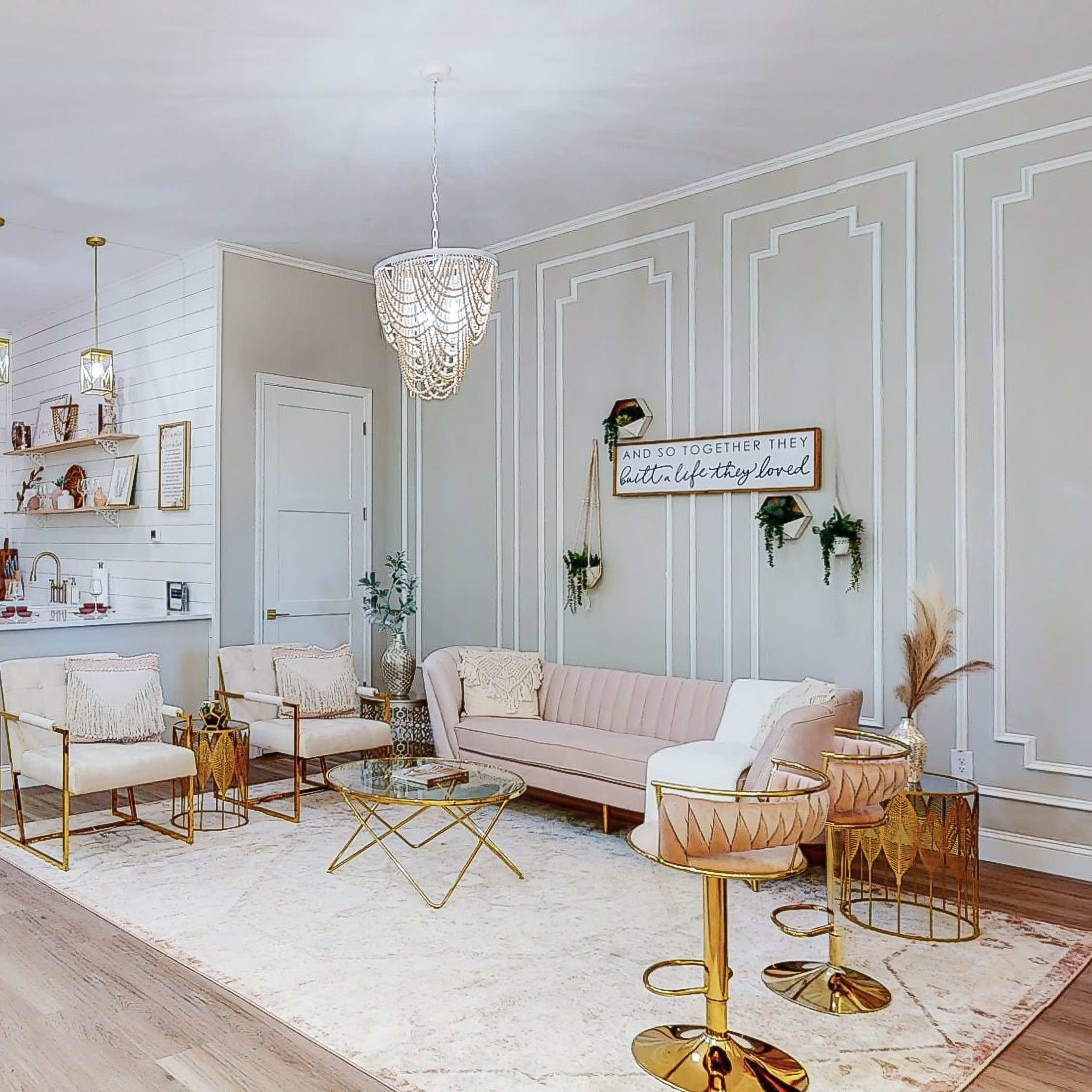 A modern, stylish living space featuring a pink sofa, elegant light fixtures, a rug, and a kitchenette with decorative wall shelves.