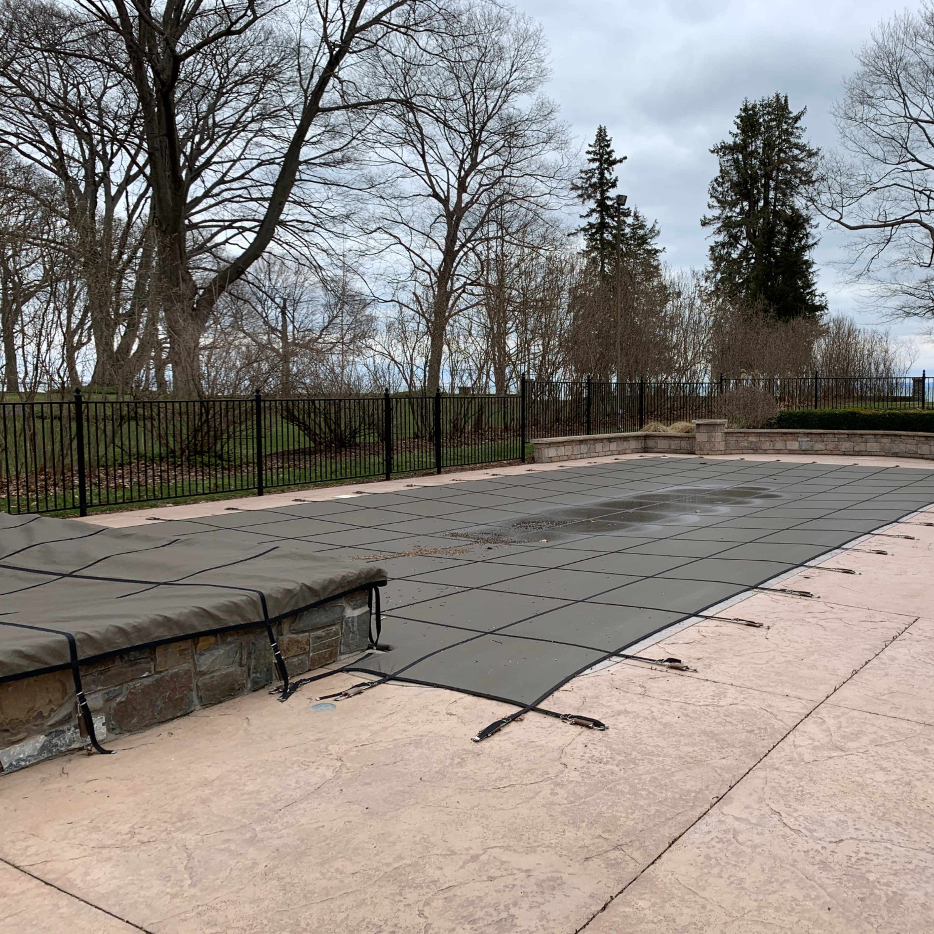 A rectangular swimming pool is covered with a tarp and surrounded by stone coping, with a fenced lawn and bare trees in the background.