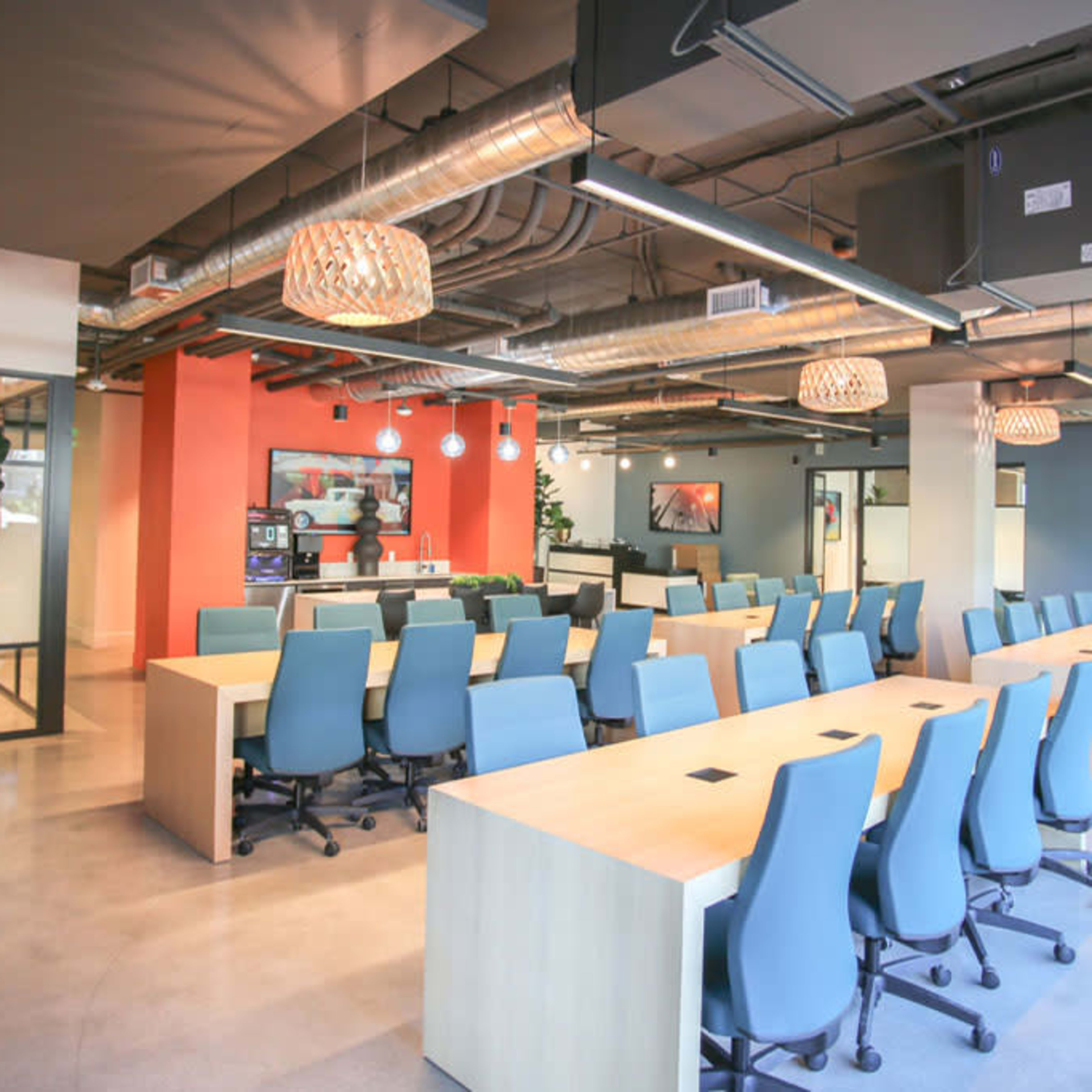 A modern office space with a long communal table surrounded by blue chairs, brightly colored walls, and hanging light fixtures.