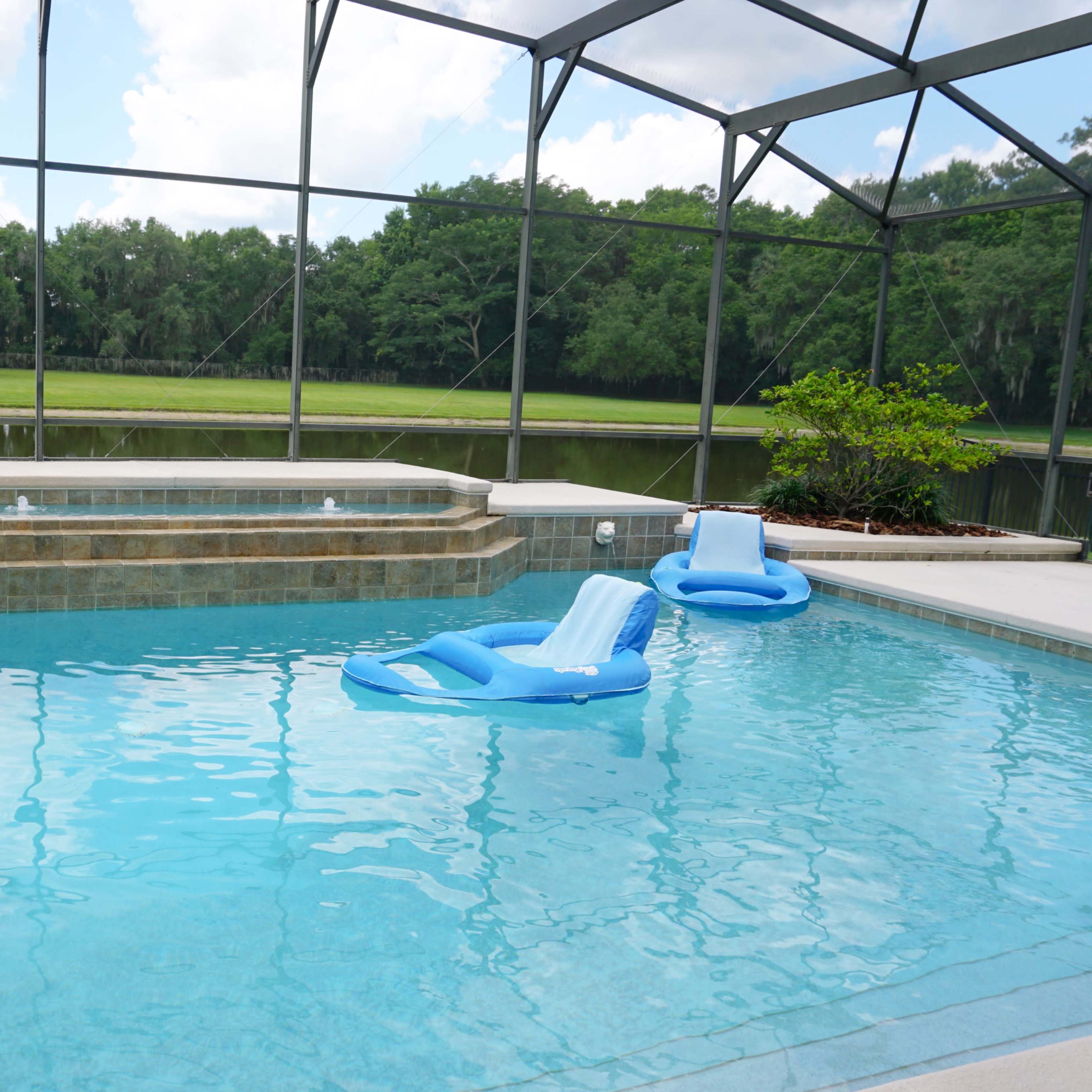 A swimming pool with two blue loungers, a raised spa area, and a view of a green landscape through a screened enclosure.