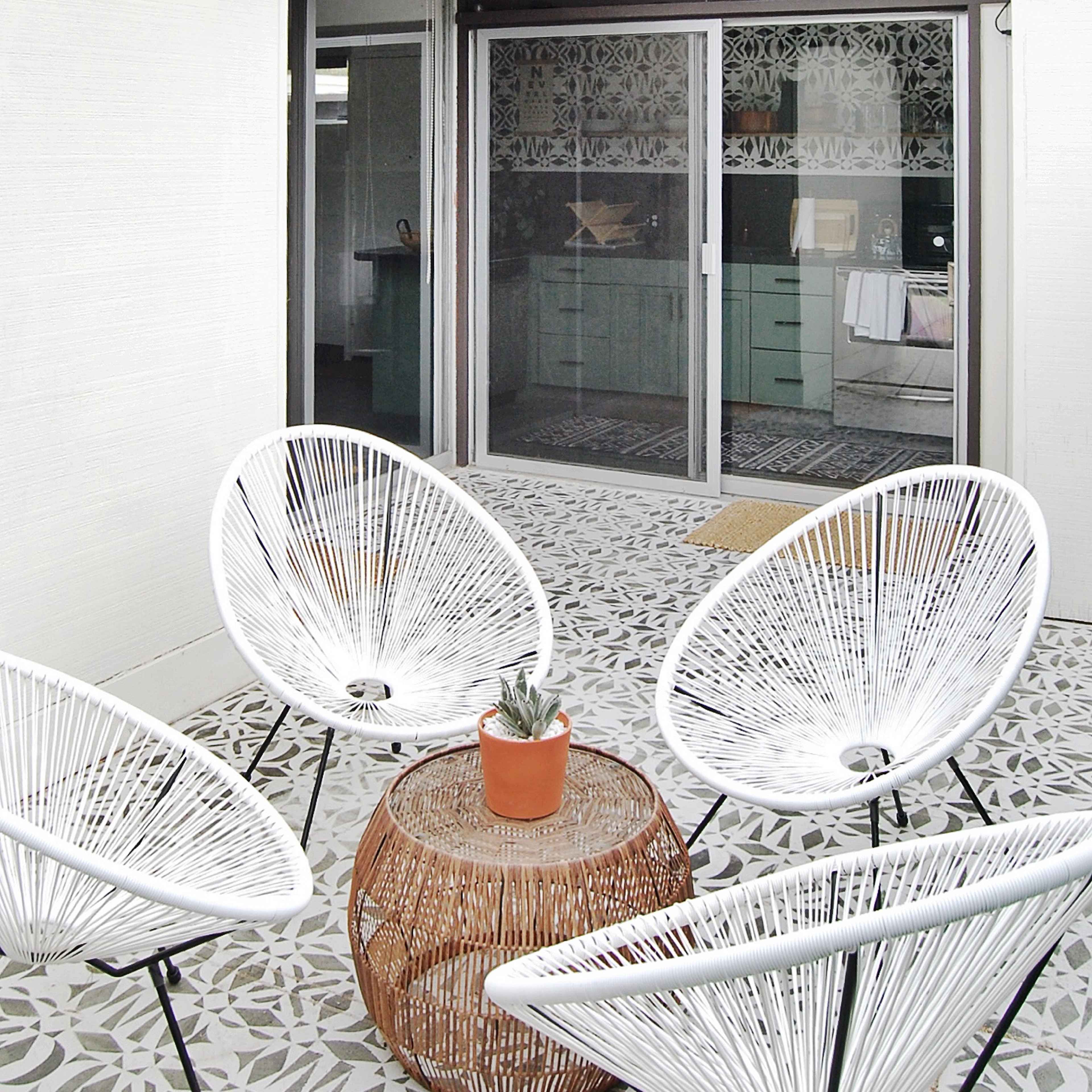 A patio features four white chairs arranged around a wooden table with a potted plant, surrounded by palm trees and string lights.