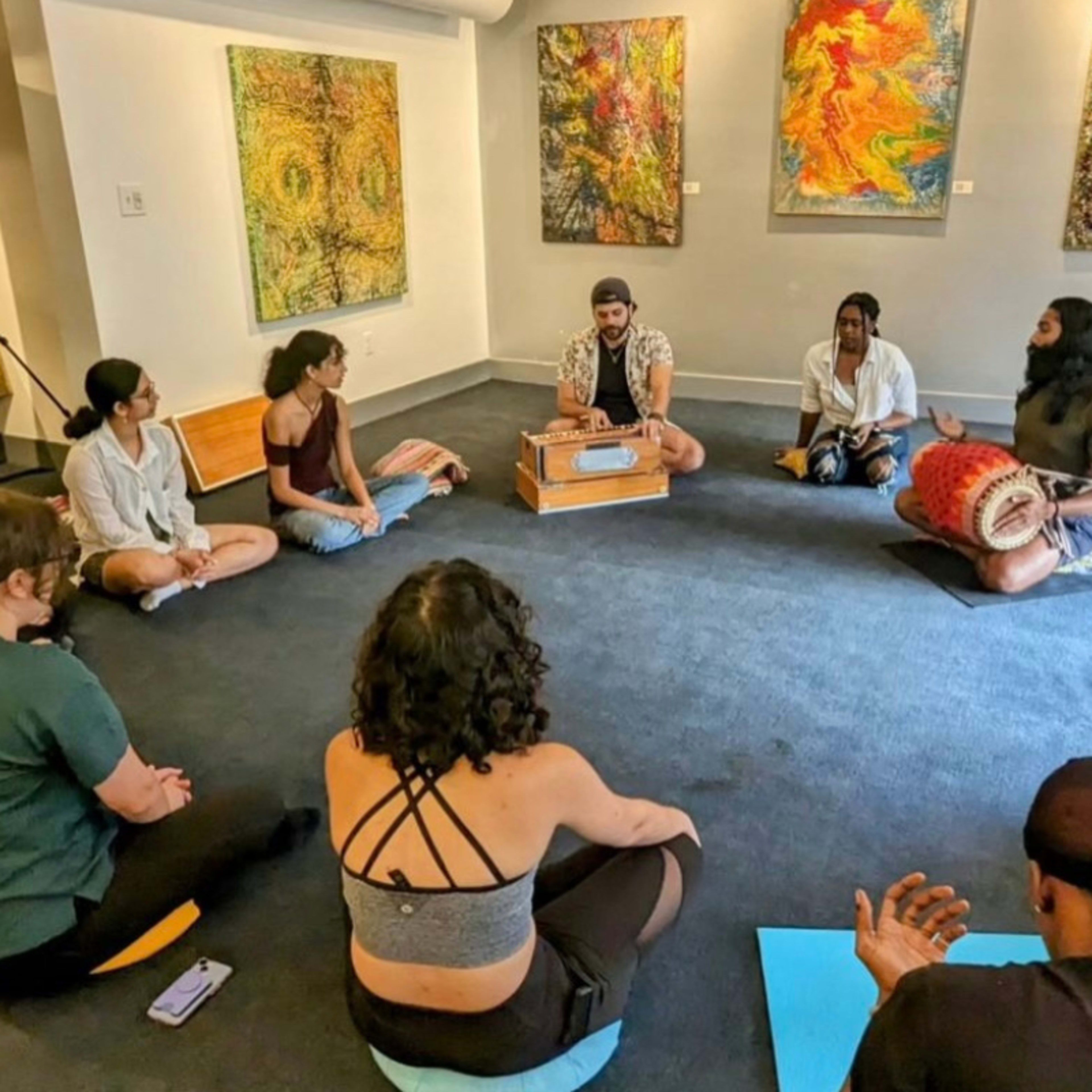 A group of people sit in a circle on the floor of an art gallery, engaging in an interactive session while surrounded by colorful abstract paintings on the walls.