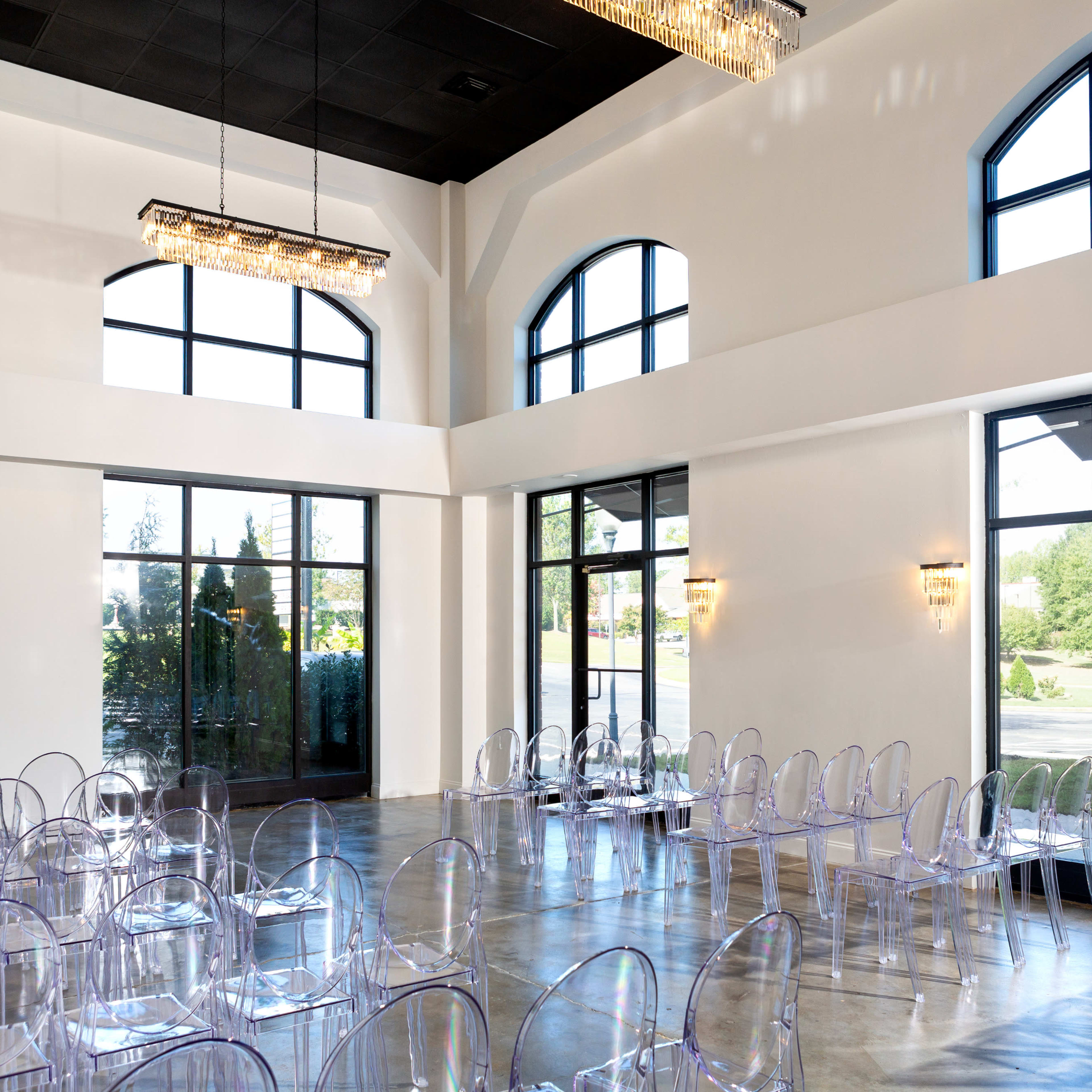 A spacious event room features clear acrylic chairs arranged neatly, large windows allowing natural light, and modern chandeliers above.