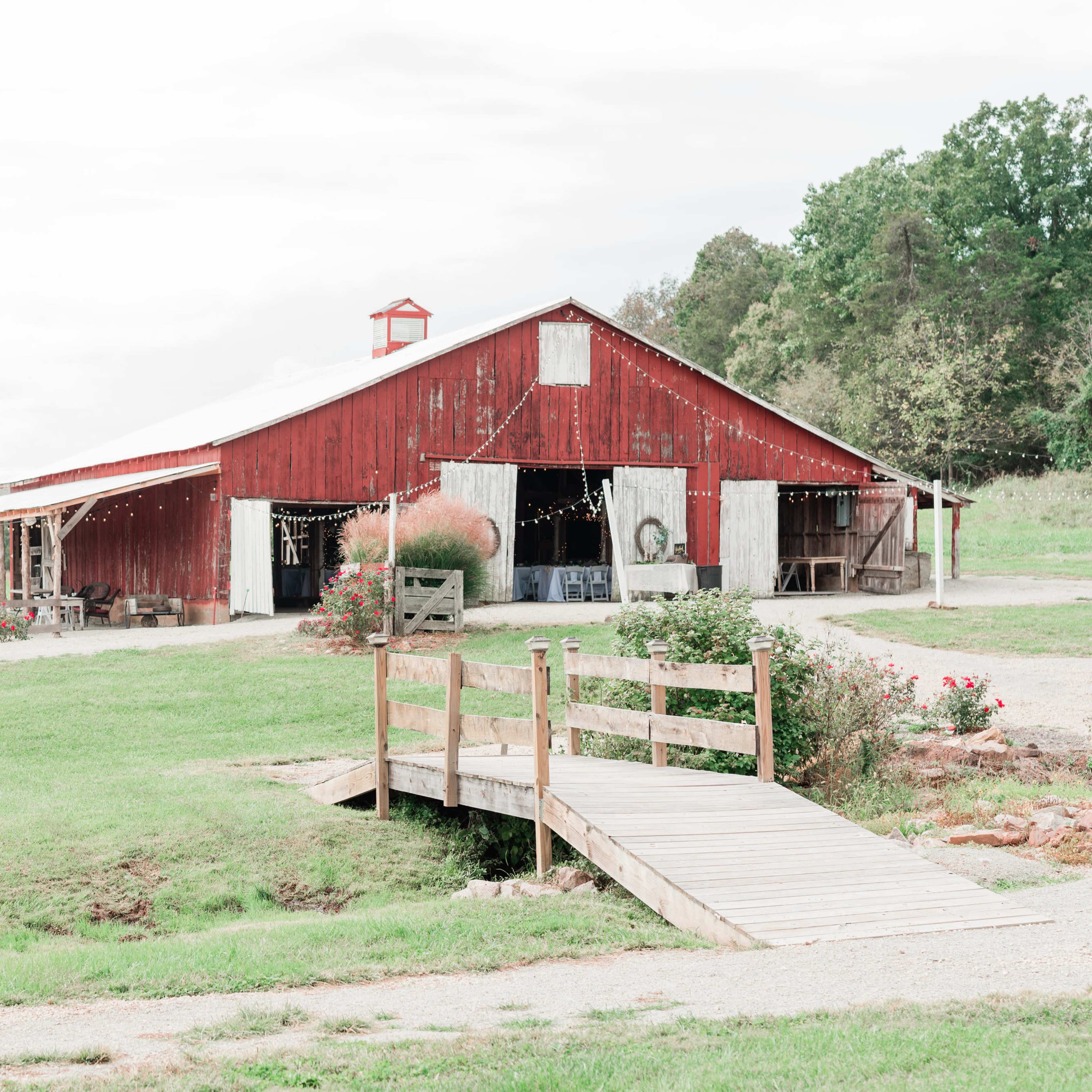 A red barn with a porch and a wooden bridge is situated on a grassy area surrounded by trees.