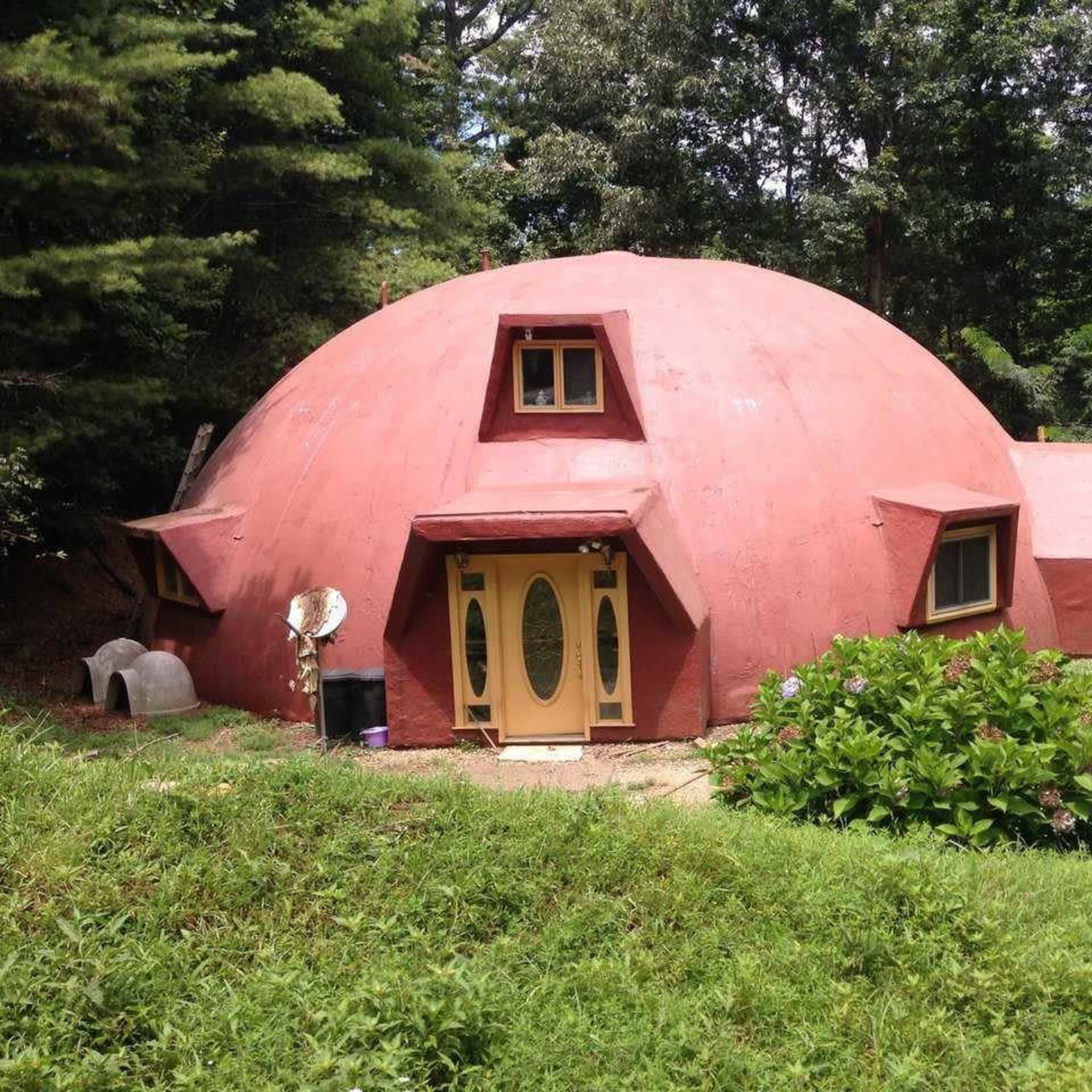 A red, dome-shaped house with a unique architectural design, set in a green, wooded area.
