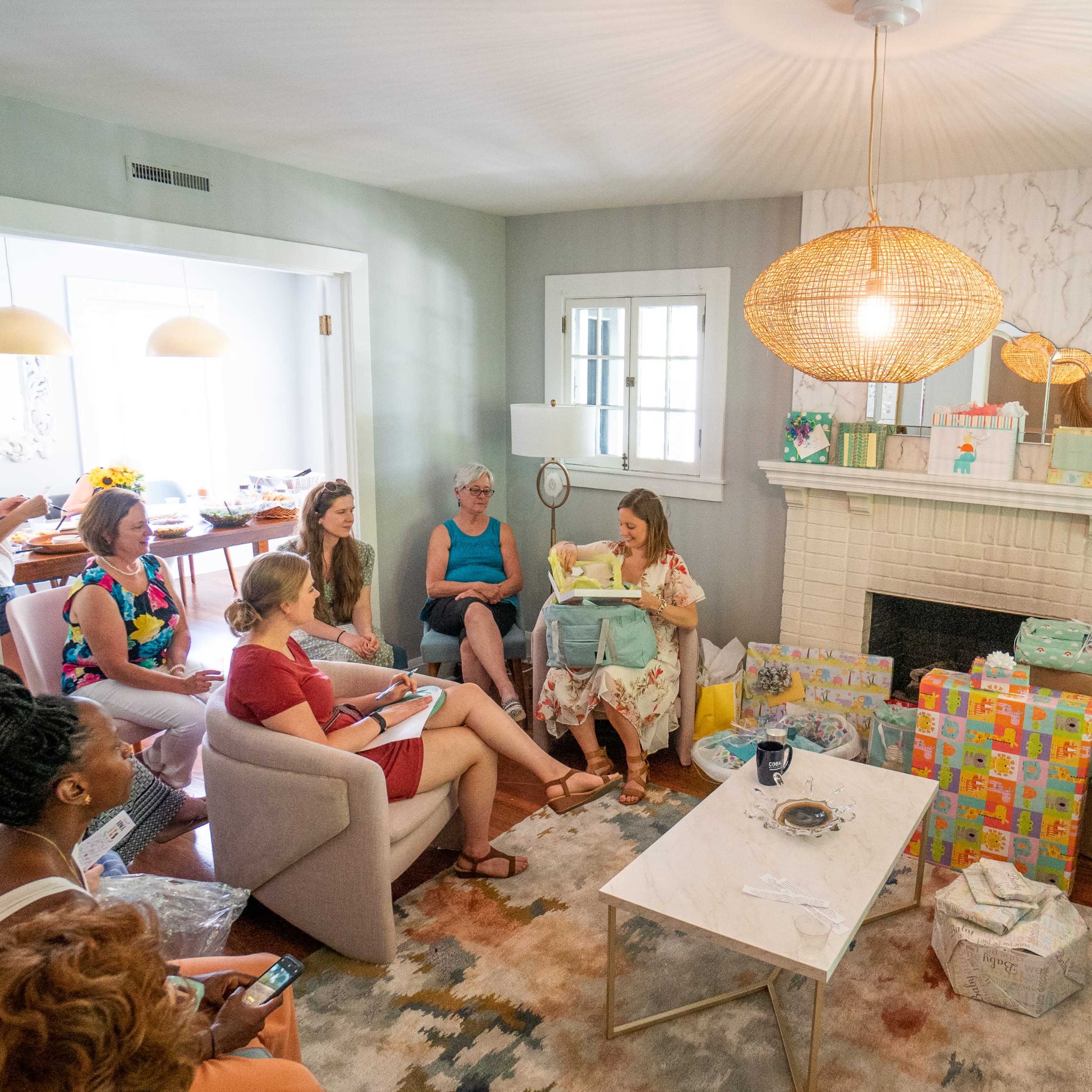 A group of women gathers in a living room around a coffee table, with gifts and packages arranged nearby, while one woman presents something to the others.