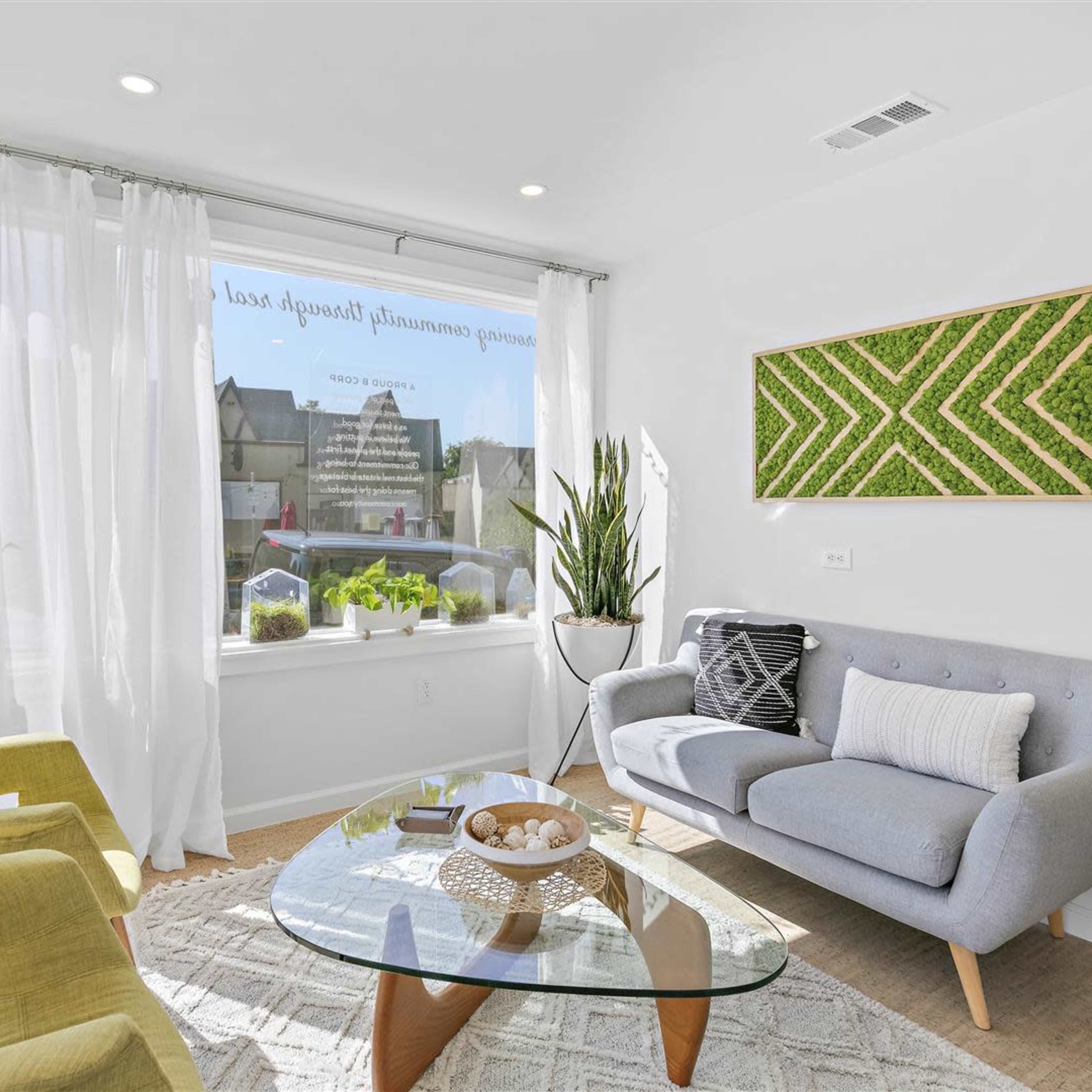 A modern waiting room features a gray sofa, two yellow chairs, a glass coffee table, and a large window with plants and natural light.