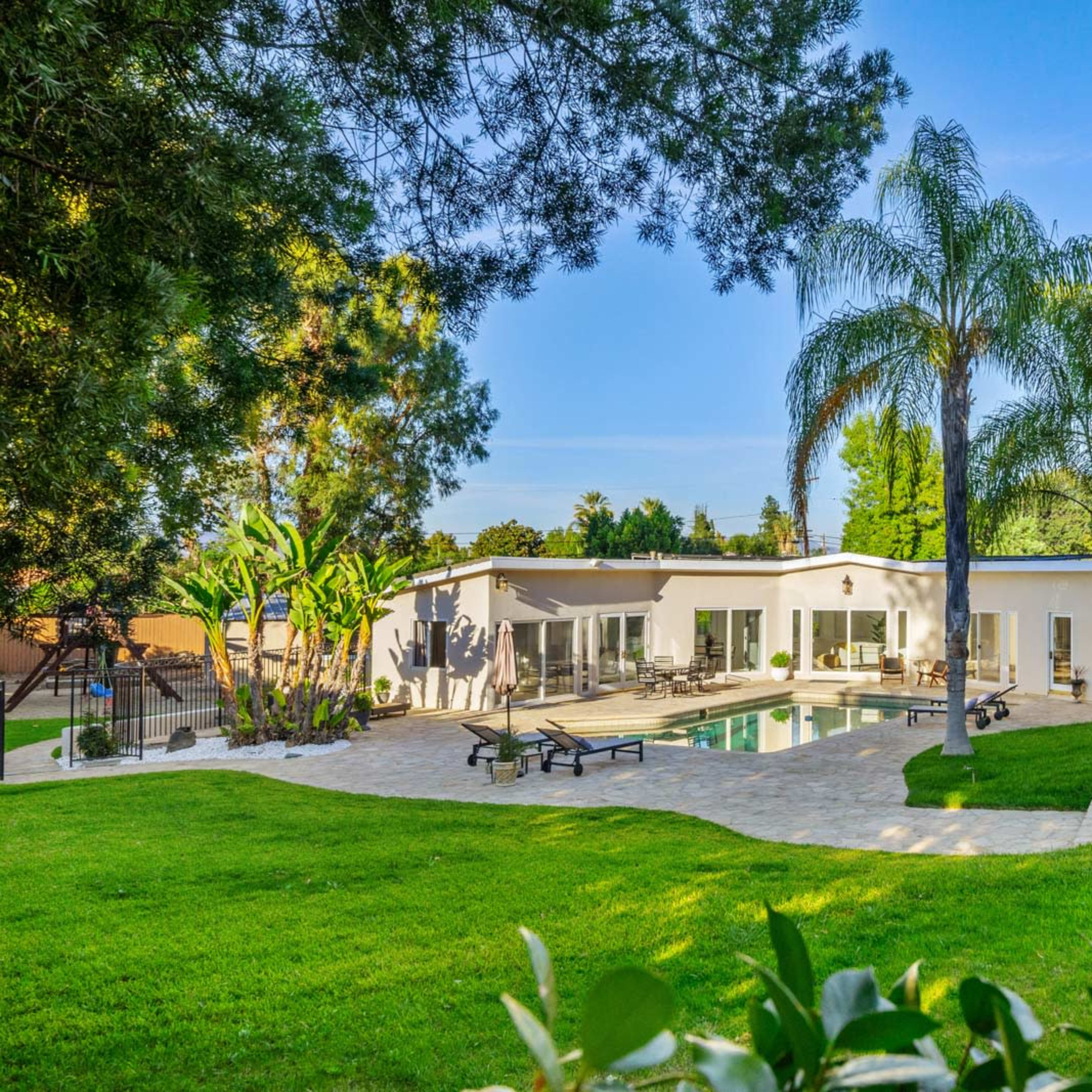 A modern house with large windows and a swimming pool surrounded by lush greenery and palm trees.
