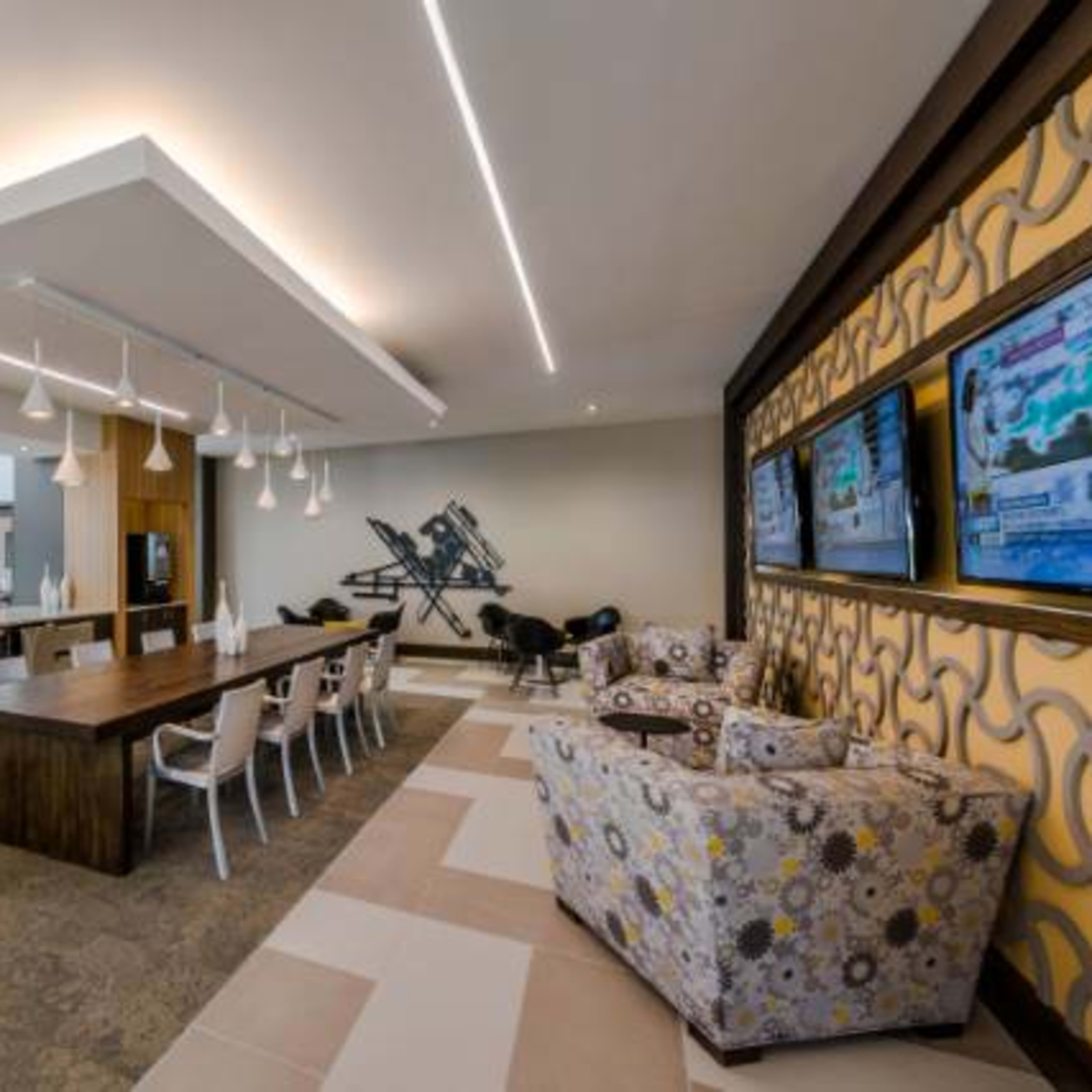 A modern dining area with a long wooden table, white chairs, and a kitchen space with stainless steel appliances, featuring television screens on the wall.