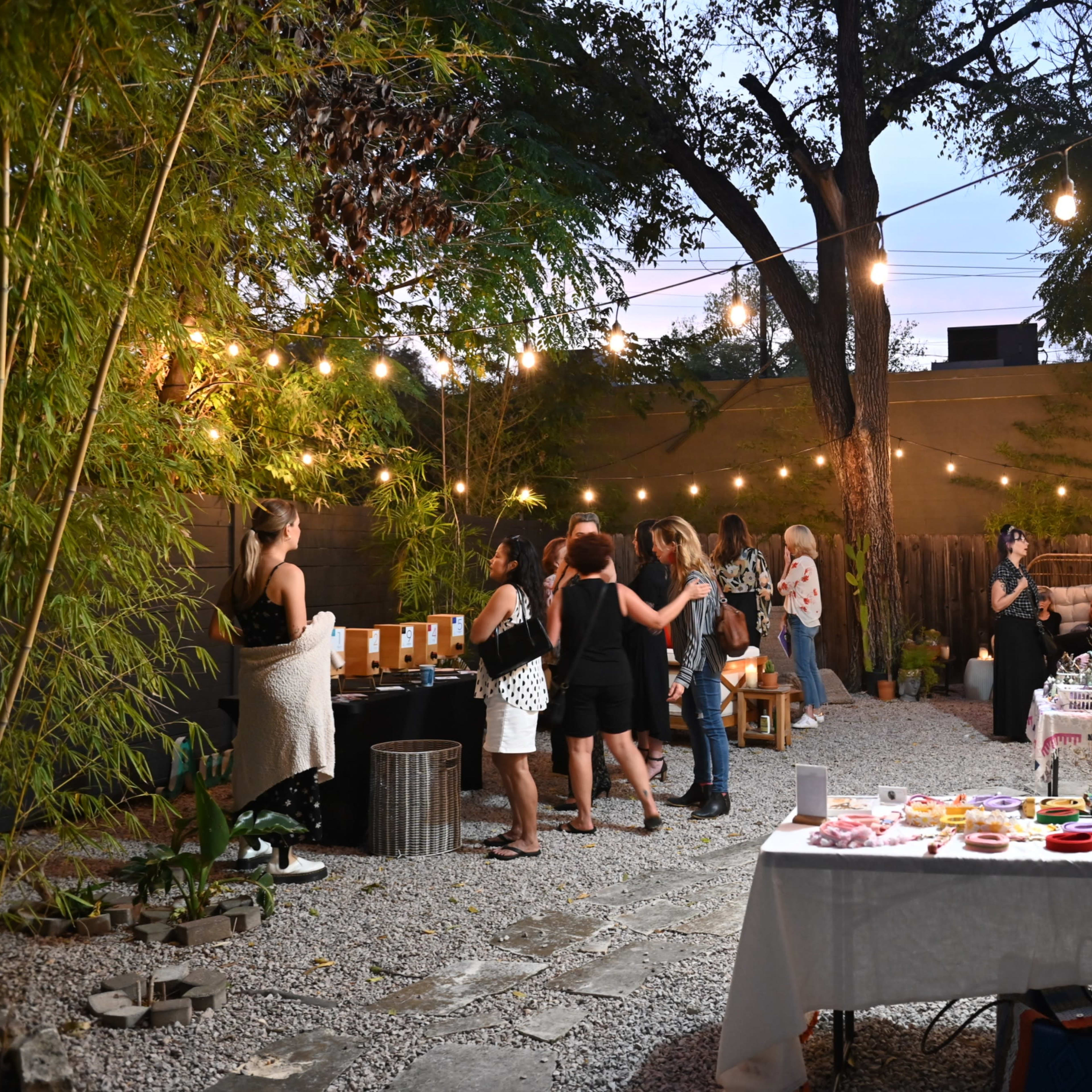 A gathering takes place in a backyard with string lights, where people socialize and various handmade goods are displayed on tables.