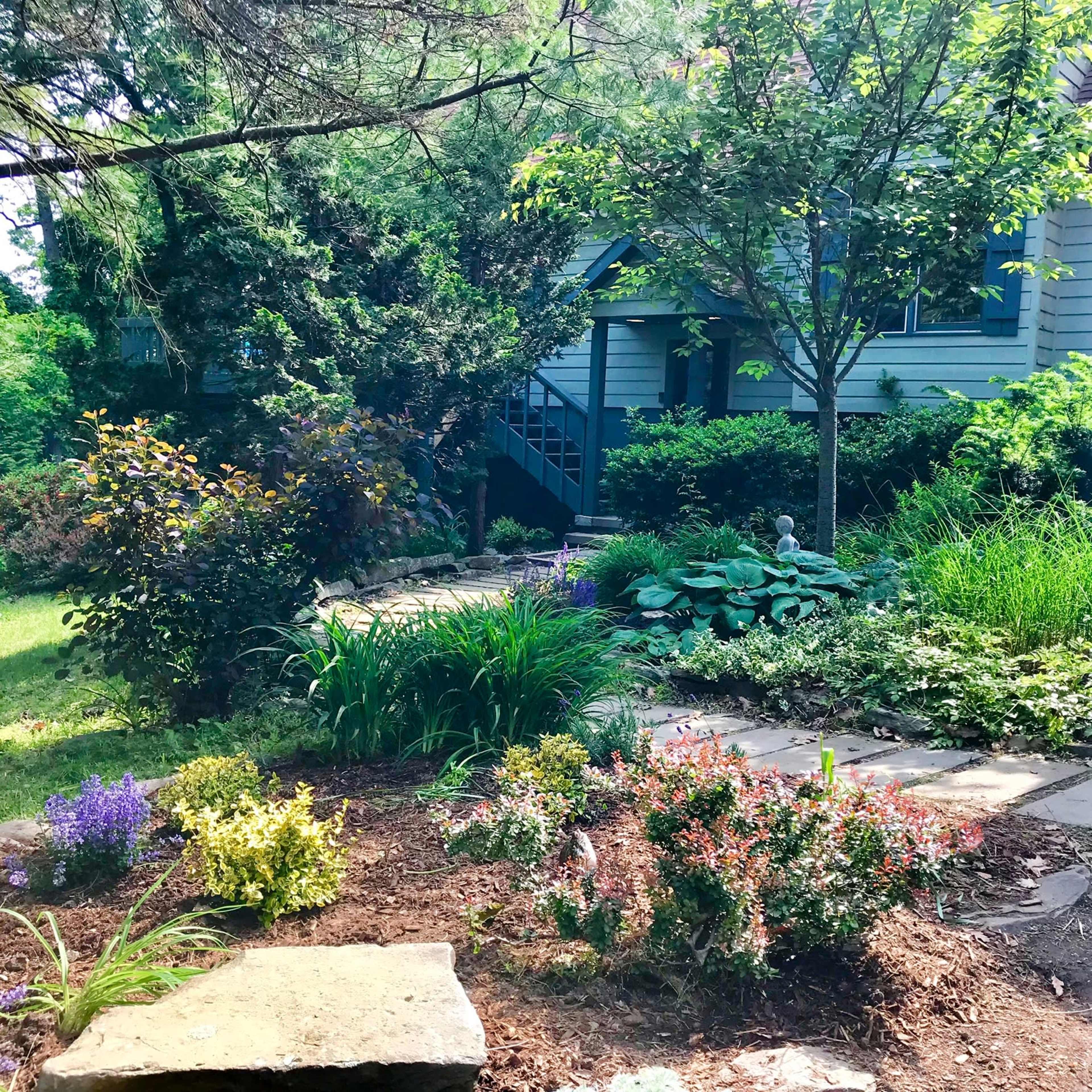 A path made of stone leads through a lush garden with various plants and a small statue in front of a house.