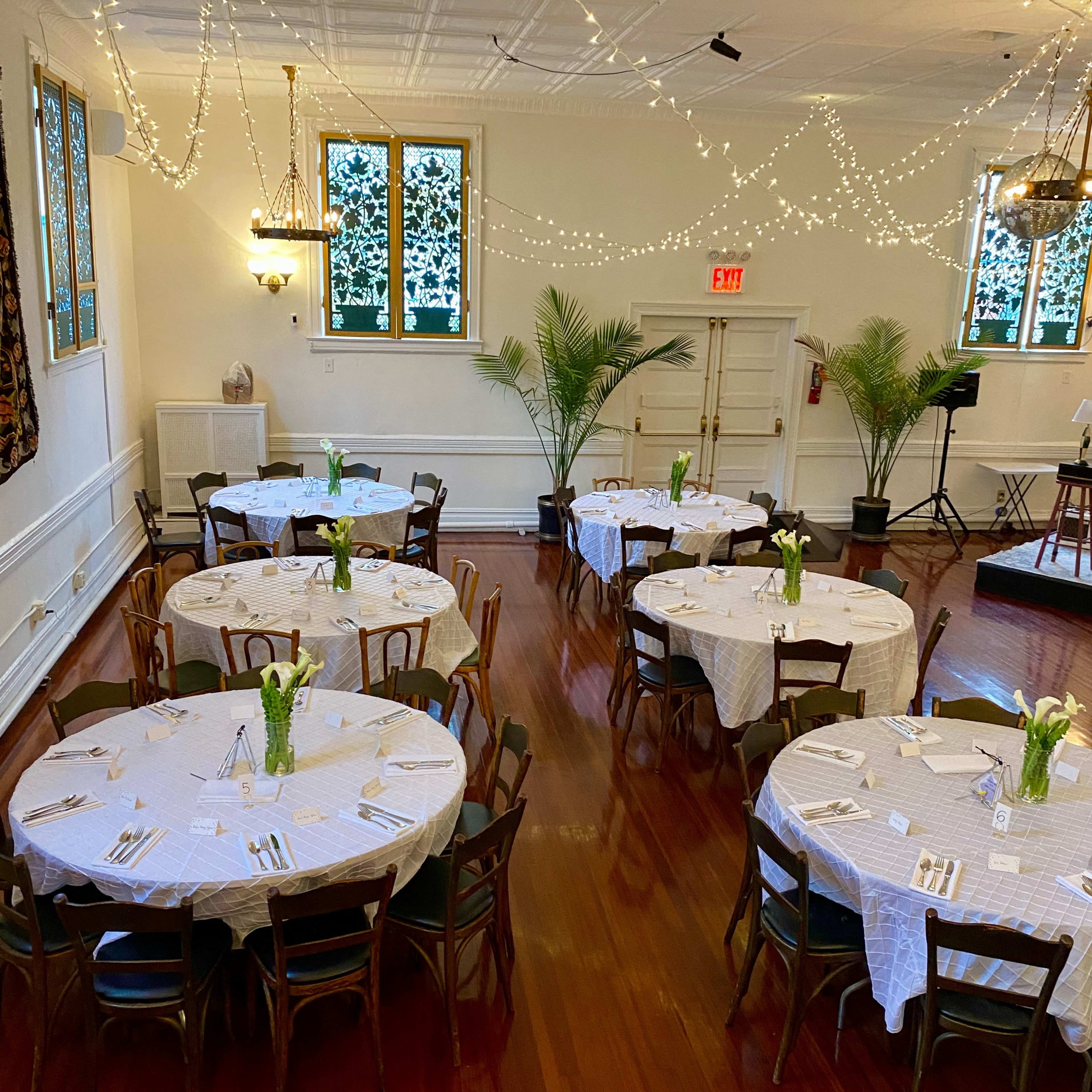A banquet hall with several round tables set with white tablecloths and centerpieces, illuminated by soft string lights and flanked by potted plants.