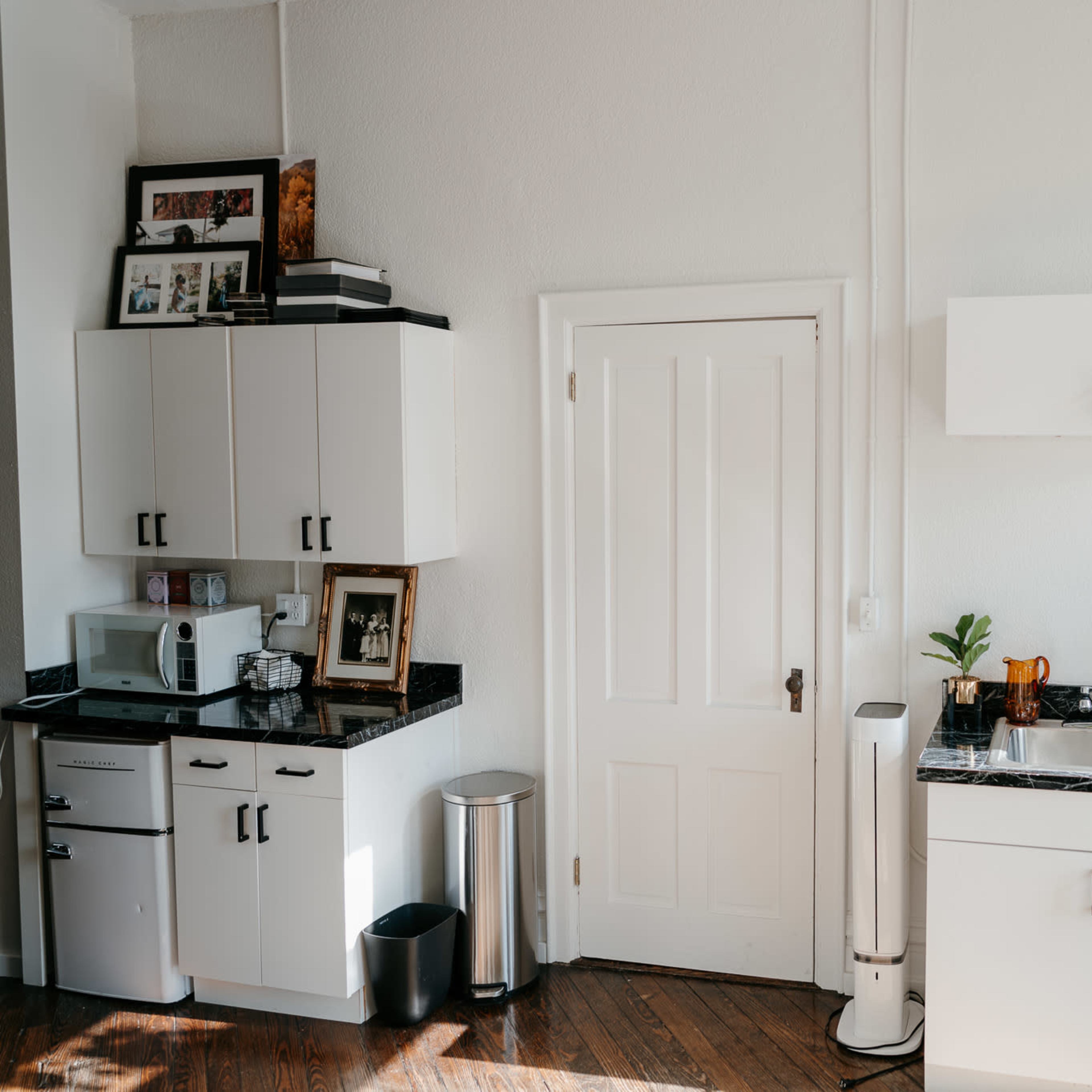 A small kitchen features white cabinets, a stainless steel sink, a microwave, and a trash can, with a door leading to another room.