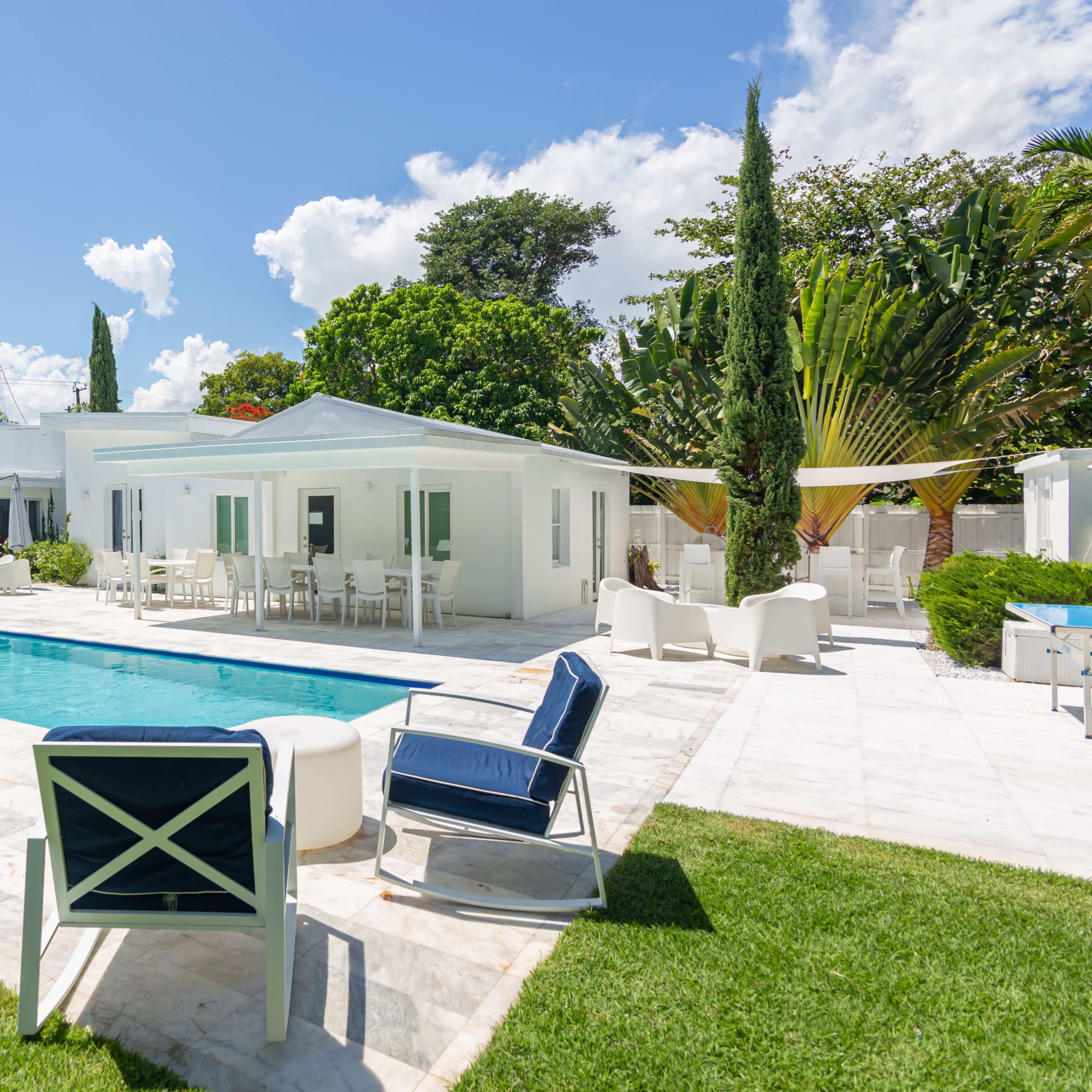 A modern backyard with a swimming pool, lounge chairs, and a white house surrounded by greenery and palm trees.