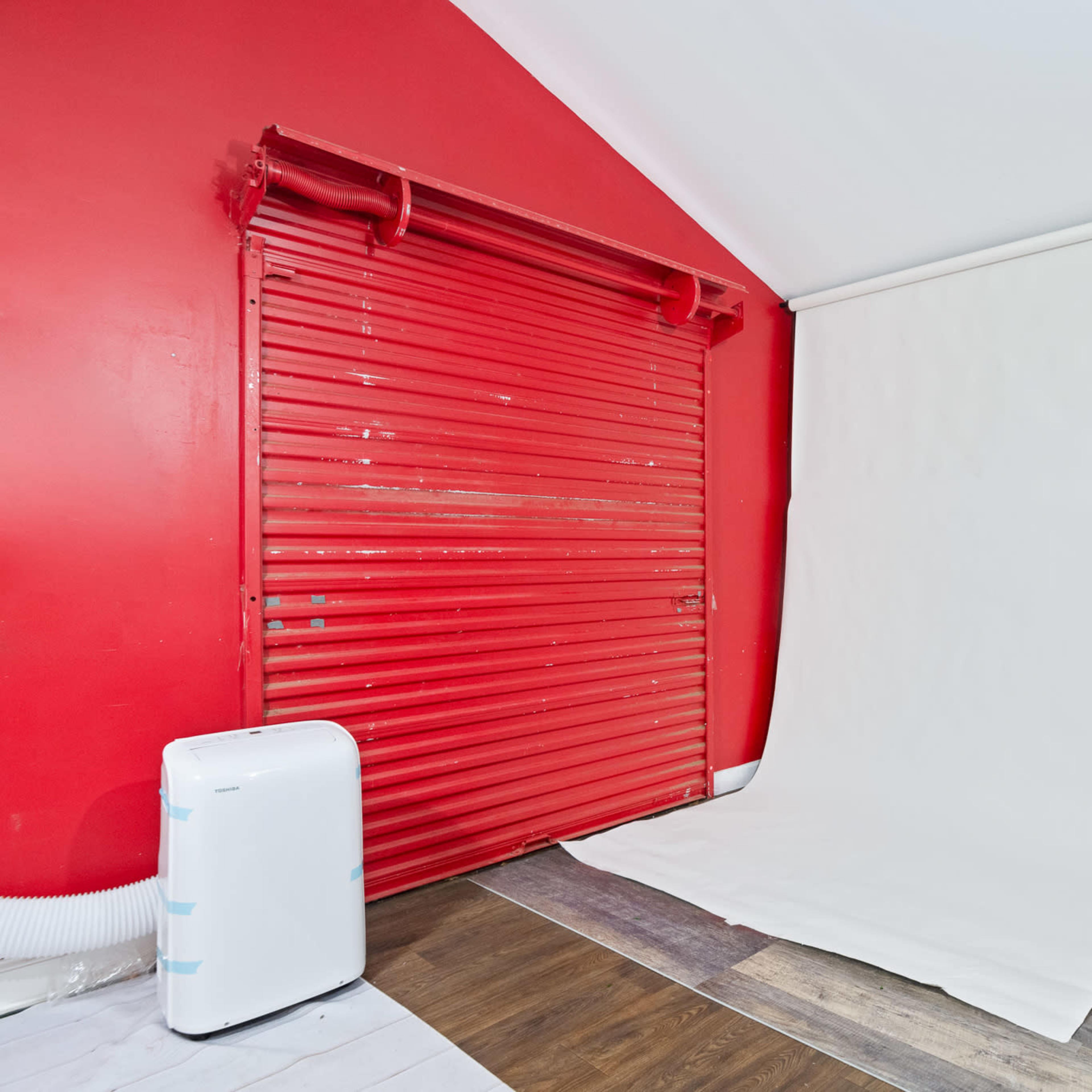A room with a red wall, a white backdrop on the floor, and a door on one side, complemented by a white appliance.