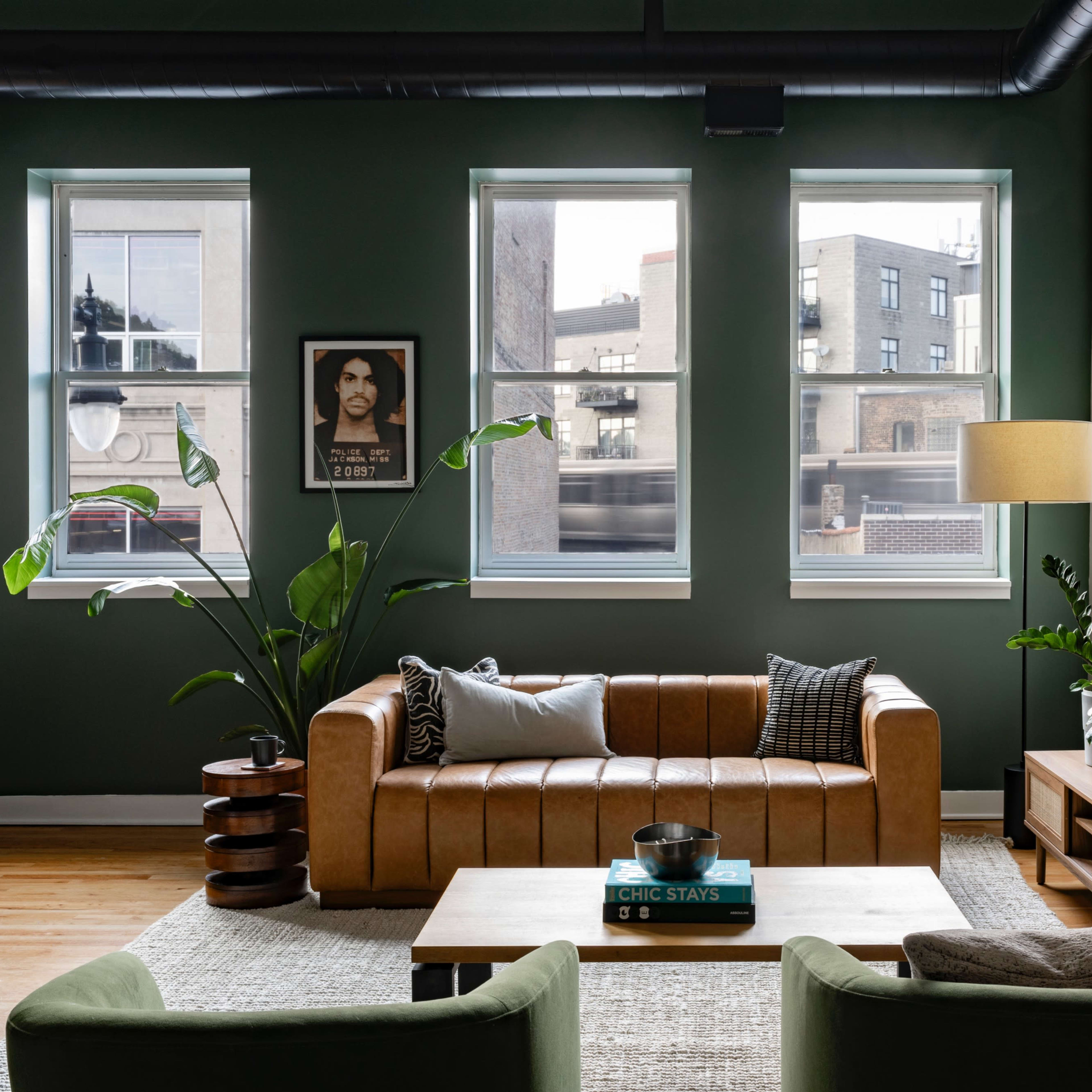 A modern living room featuring a brown leather sofa, a green accent wall, large windows, and a television mounted on the wall.