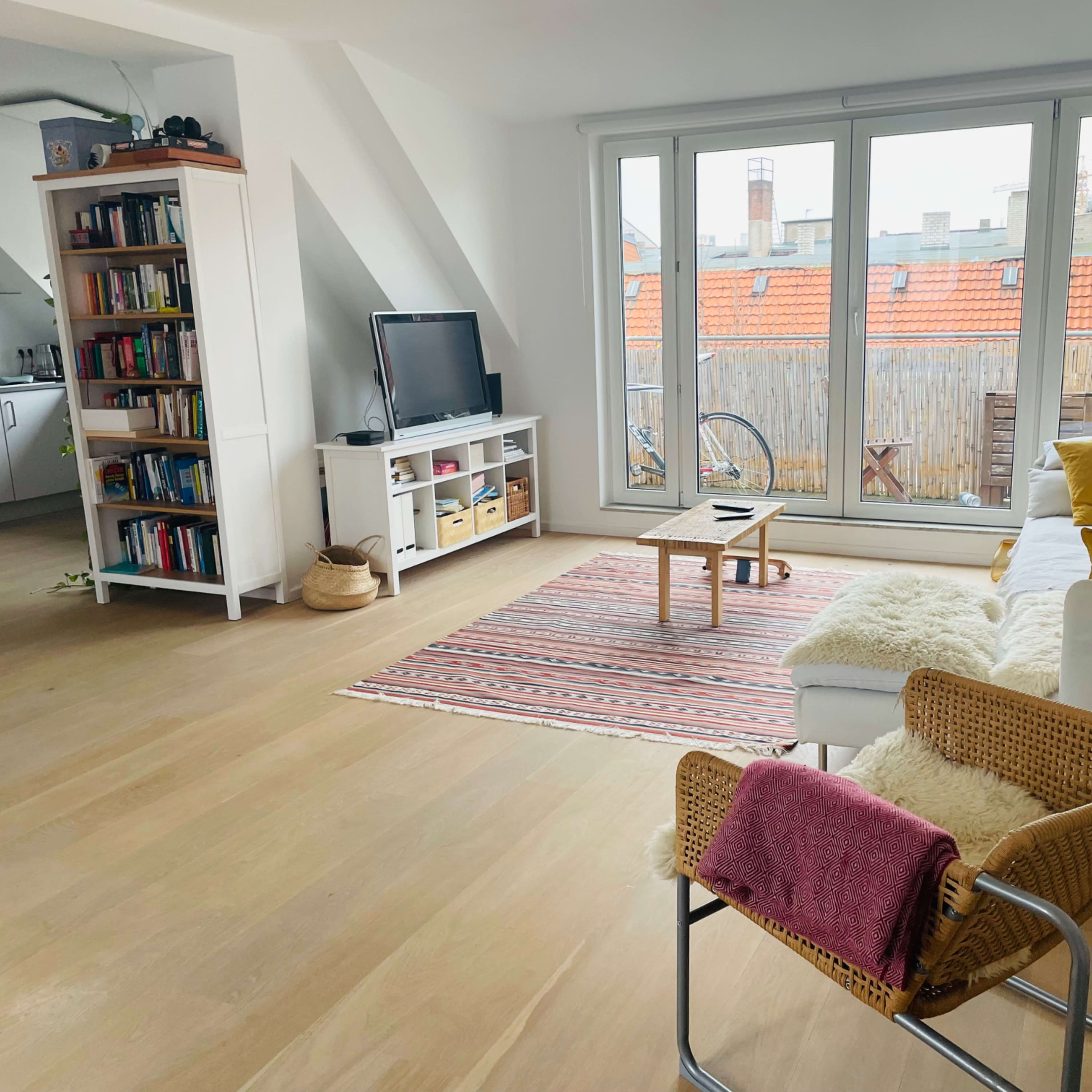 A bright living room features a white sofa, a coffee table on a patterned rug, a bookshelf filled with books, and large windows overlooking a courtyard.