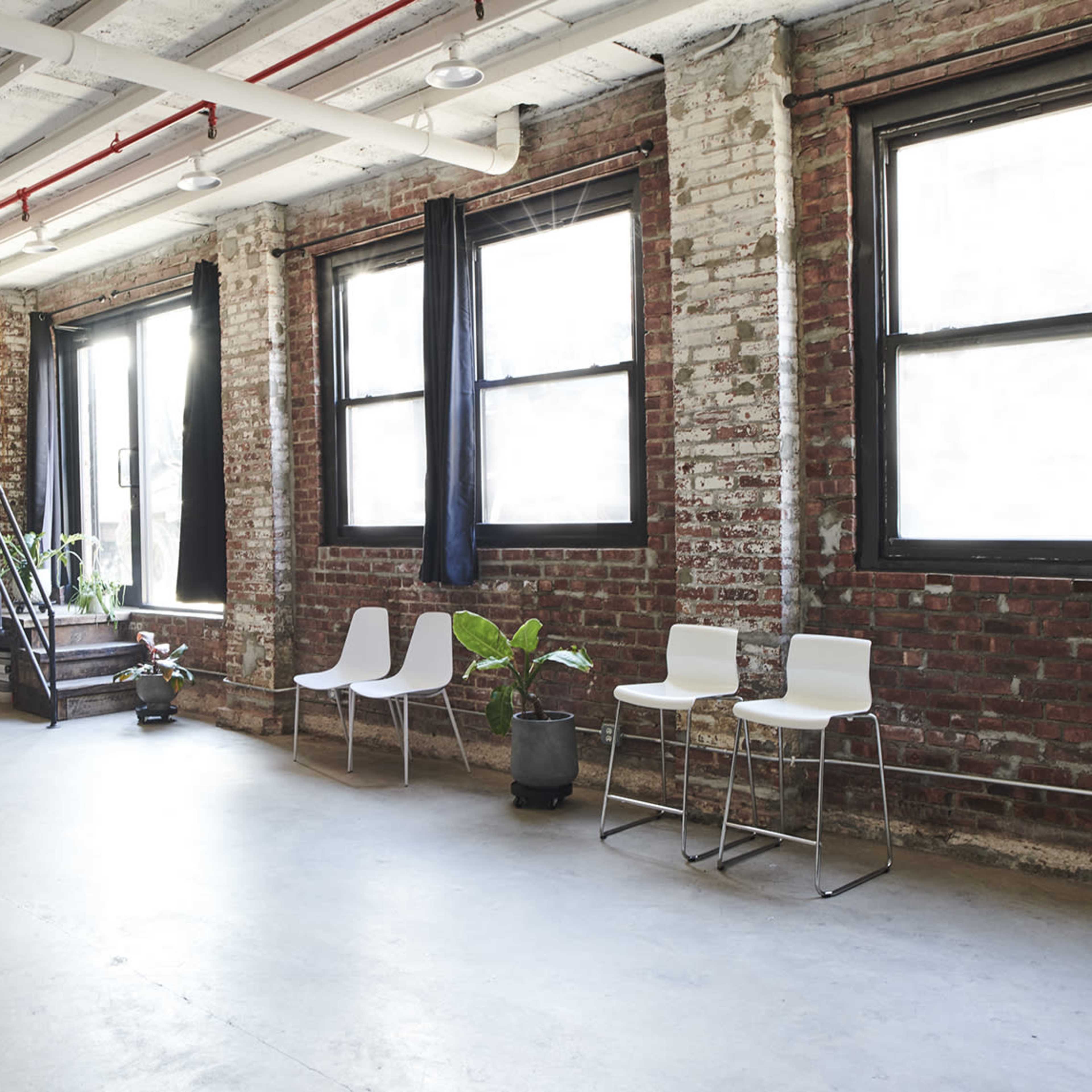 An industrial-style interior with exposed brick walls, large windows, and several white chairs arranged along one side.