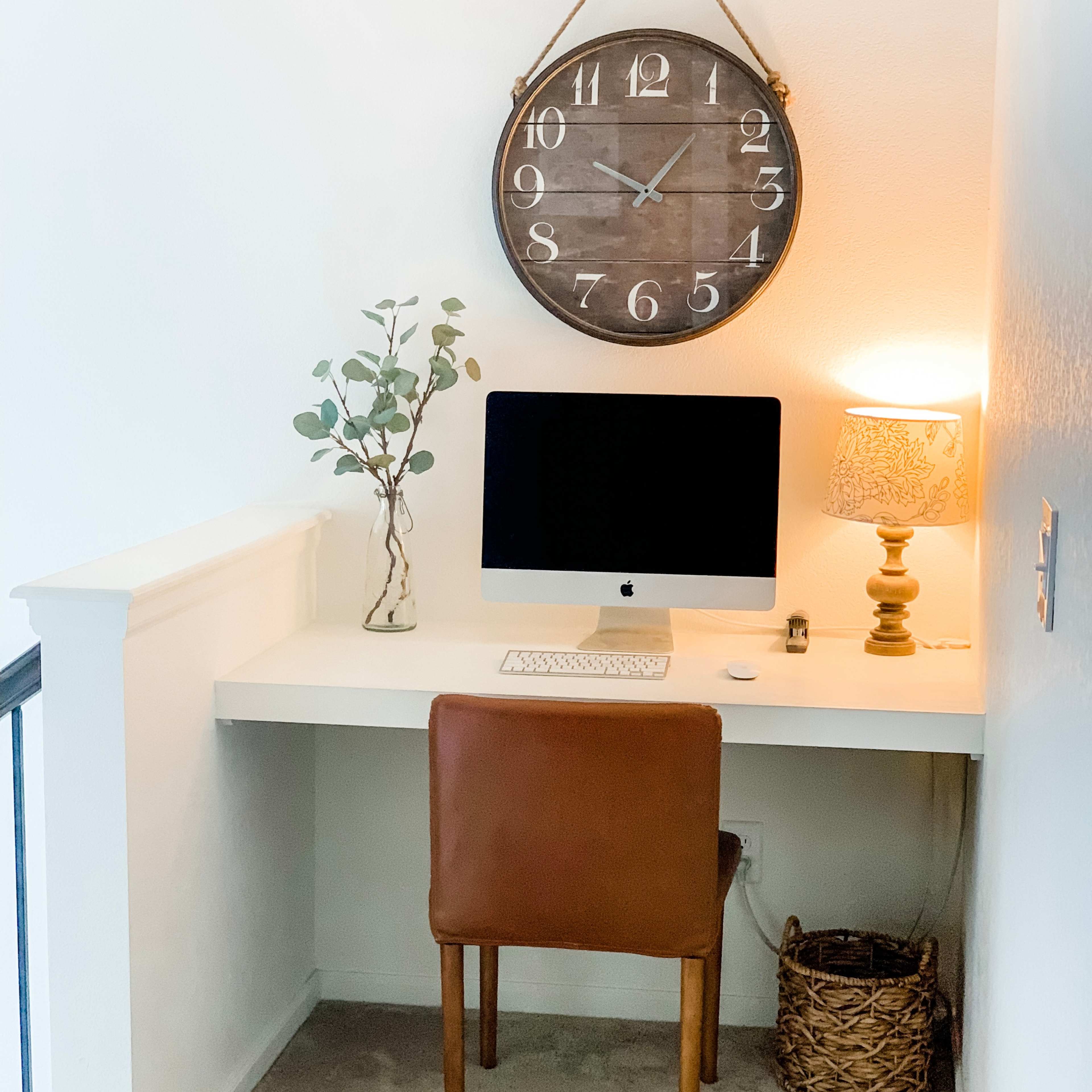 A minimalist workspace features an iMac on a white desk, accompanied by a brown chair, decorative clock, lamp, and a plant.