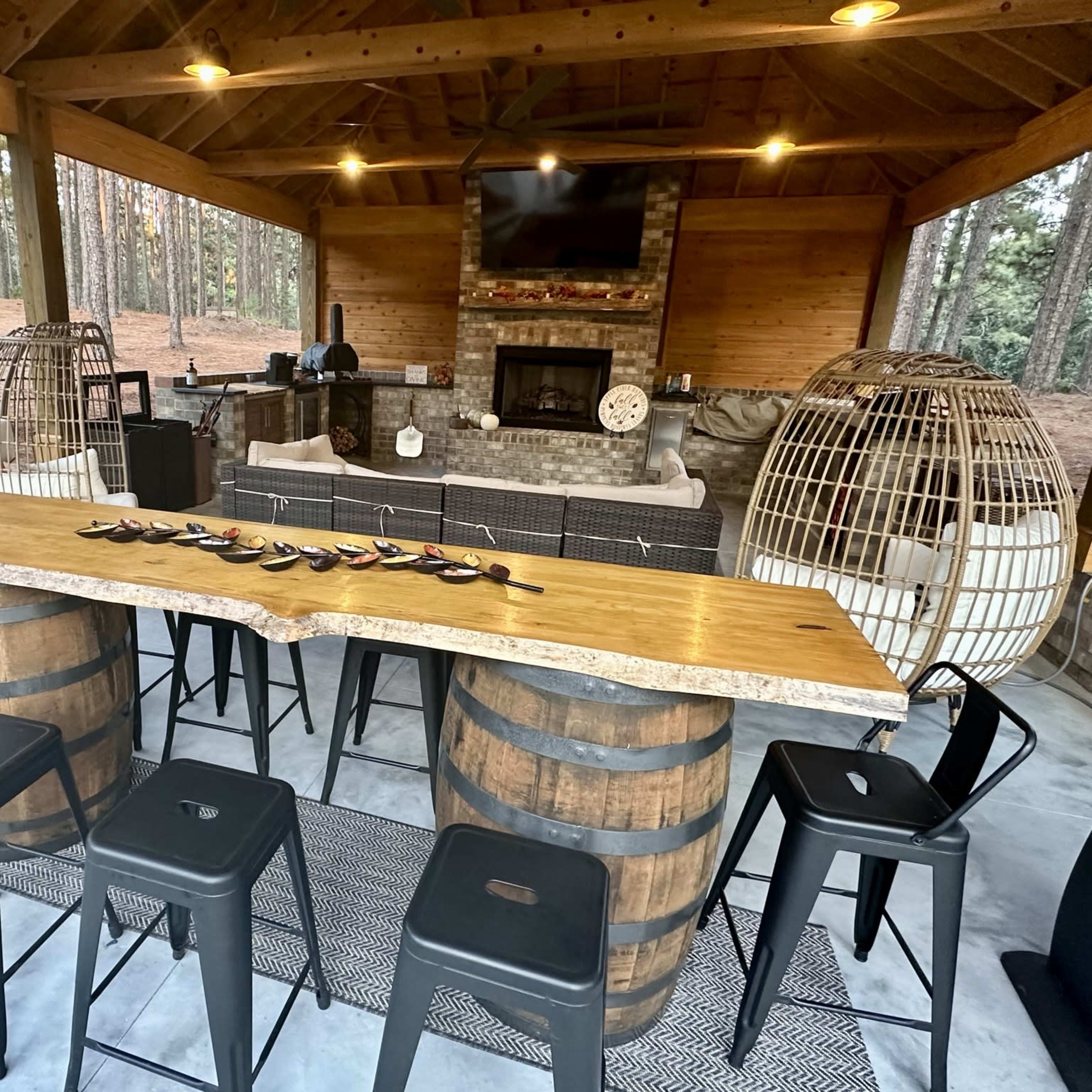An outdoor bar area with a wooden table supported by barrel bases, surrounded by metal stools, and a stone fireplace in the background.