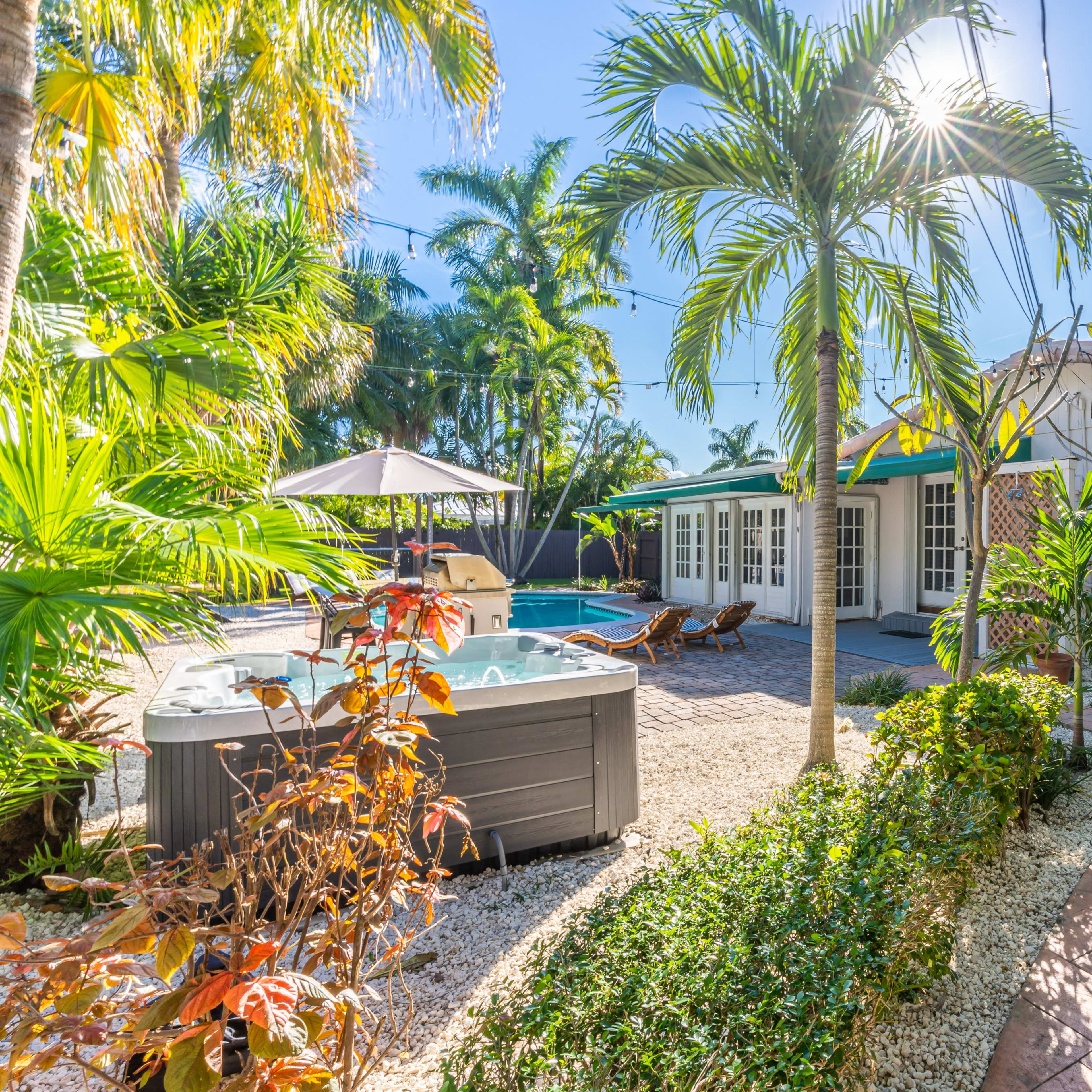 A landscaped tropical backyard featuring a hot tub, a pool, and a white house with large windows surrounded by palm trees and shrubs.