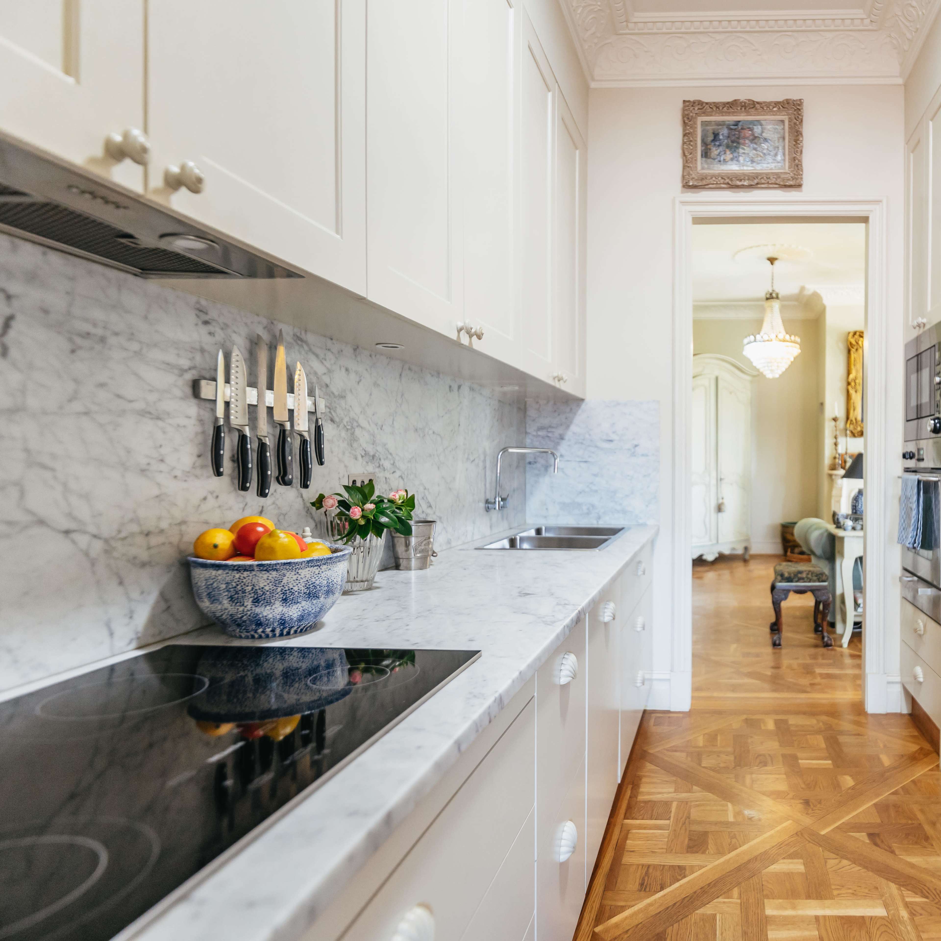 A narrow kitchen features marble countertops, a row of knives on the wall, and cabinets filled with dishes, leading to a partially visible living area.