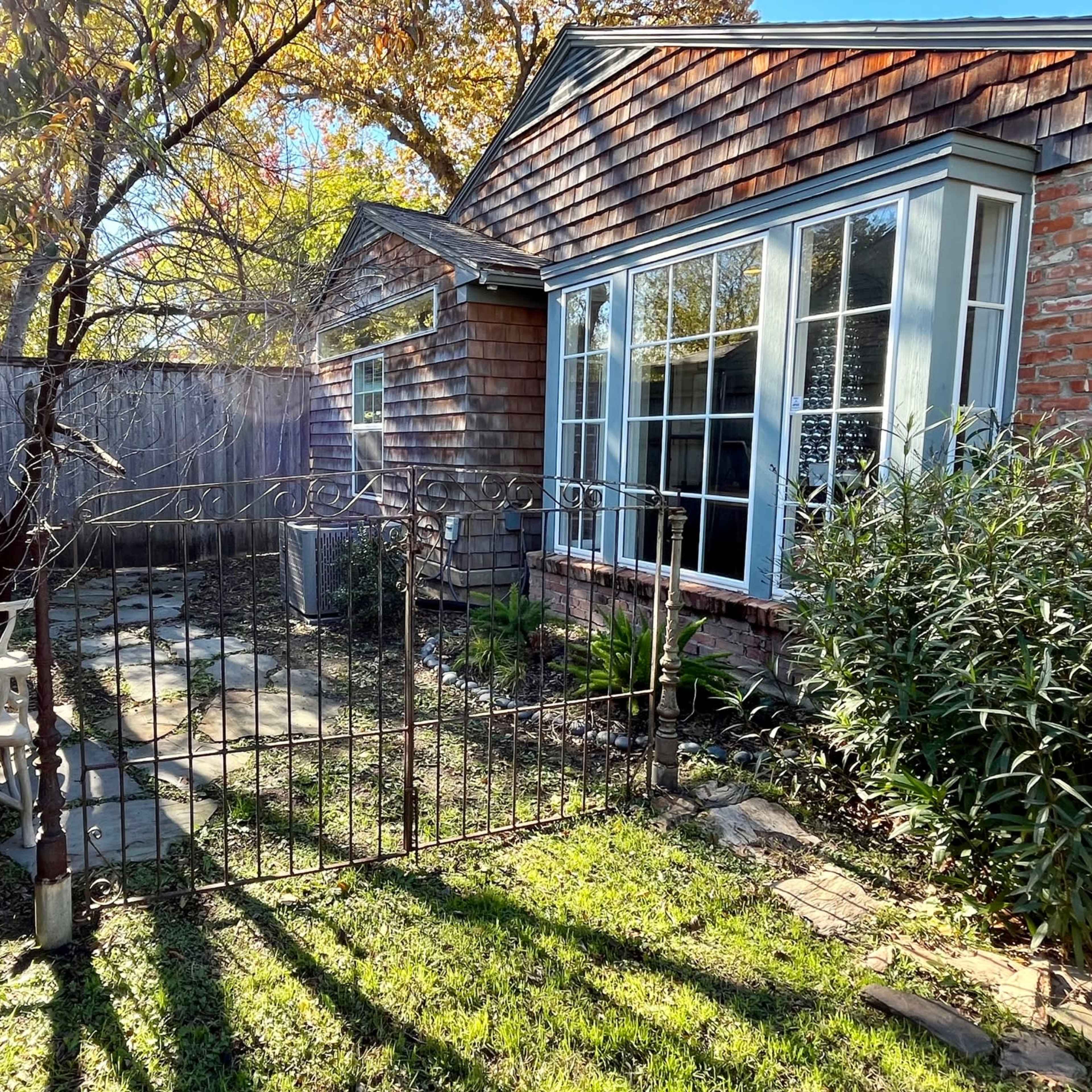 A small courtyard features a wrought iron gate, a stone pathway, a wooden house with shingle siding, and a bench beneath a tree.